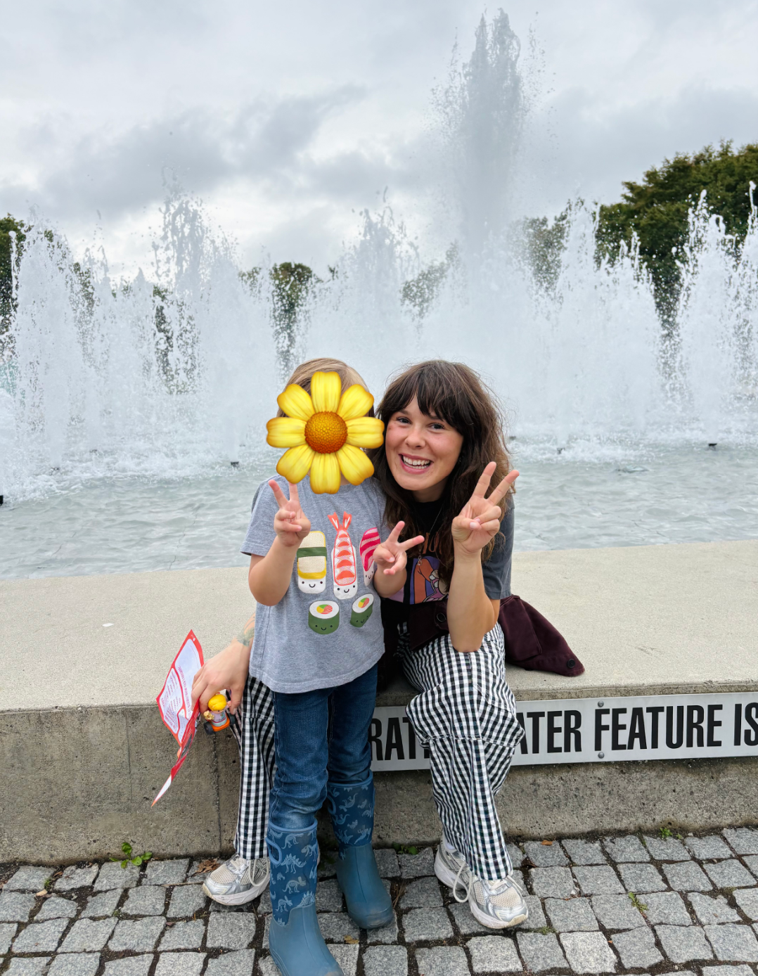 Cayley and a child making peace signs in front of a fountain on an overcast day.