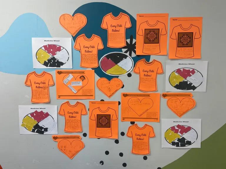 A classroom wall display of children's orange "Every Child Matters" shirt cut-outs, paper hearts, and medicine wheel worksheets.