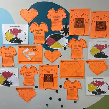 A classroom wall display of children's orange "Every Child Matters" shirt cut-outs, paper hearts, and medicine wheel worksheets.