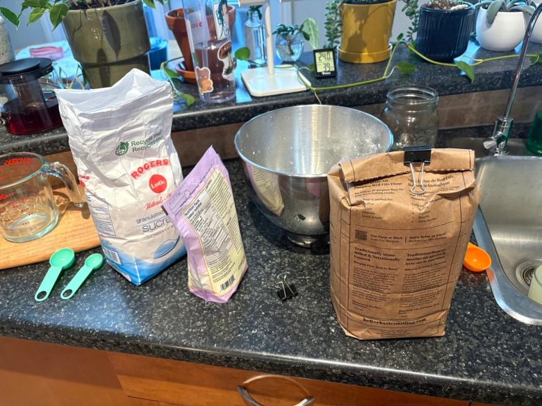 Baking ingredients set out on a kitchen counter: a bag of granulated sugar, a paper bag of stone-milled Red Fife flour, a glass measuring cup, green measuring spoons and a stainless steel mixing bowl.