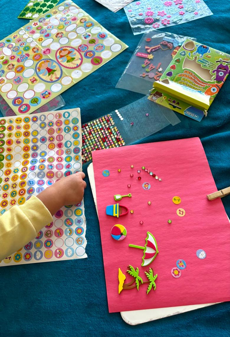 A child’s hand reaching for a sticker sheet, beside a pink page where beach-themed foam stickers – a bucket and spade, a beach ball, a parasol and palm trees – have been grouped together alongside a row of gemstone stickers.