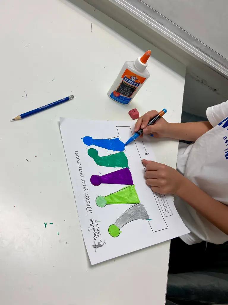 A child painting a "Design your own crown" activity sheet from The Paper Bag Princess in blue, green and purple.