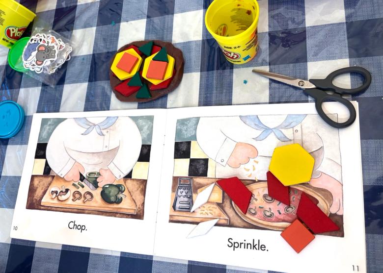 A pretend pizza made from brown Play-Doh topped with coloured geometric shape tiles, laid out on a checked tablecloth beside an open Early Reader picture book showing a chef chopping vegetables.