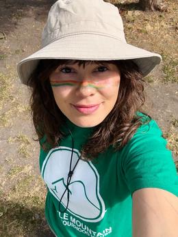 Cayley outdoors in a wide-brimmed sun hat and a green community t-shirt, with a rainbow painted across both cheeks.