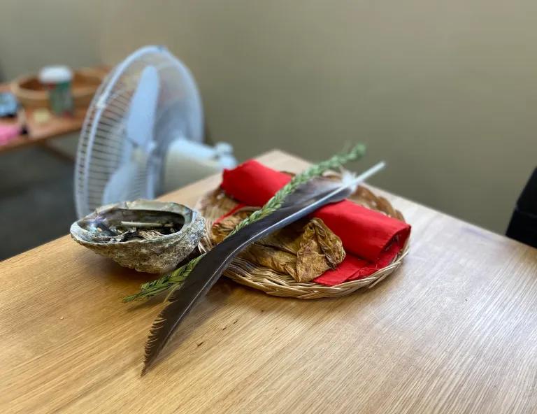 A smudging bundle resting on a classroom table: an abalone shell, dried medicines wrapped in red cloth in a woven basket, sweetgrass, and a feather.