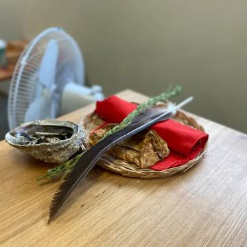 A smudging bundle resting on a classroom table: an abalone shell, dried medicines wrapped in red cloth in a woven basket, sweetgrass, and a feather.