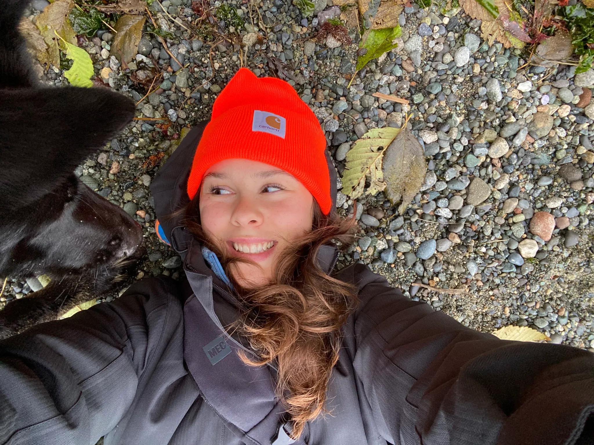 Cayley lying on a pebbled beach in an orange toque and rain jacket, laughing, with a black dog leaning into the frame.