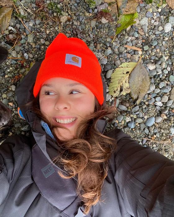 Cayley lying on a pebbled beach in an orange toque and rain jacket, laughing, with a black dog leaning into the frame.