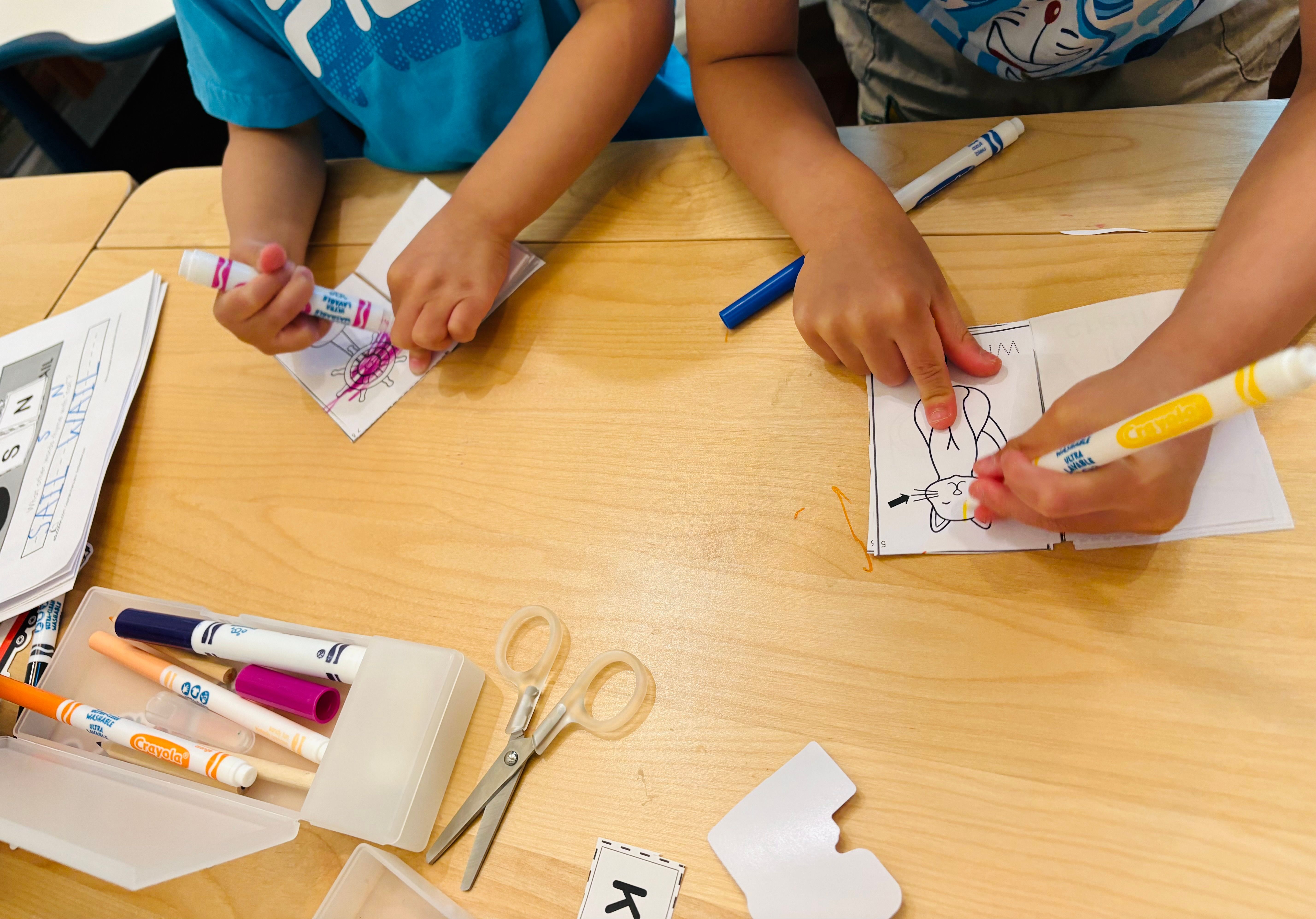 Two children colouring paper puppets with felt markers at a low table, scissors and letter cards beside them.