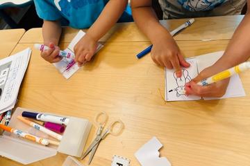 Two children colouring paper puppets with felt markers at a low table, scissors and letter cards beside them.