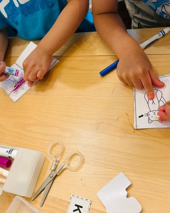 Two children colouring paper puppets with felt markers at a low table, scissors and letter cards beside them.