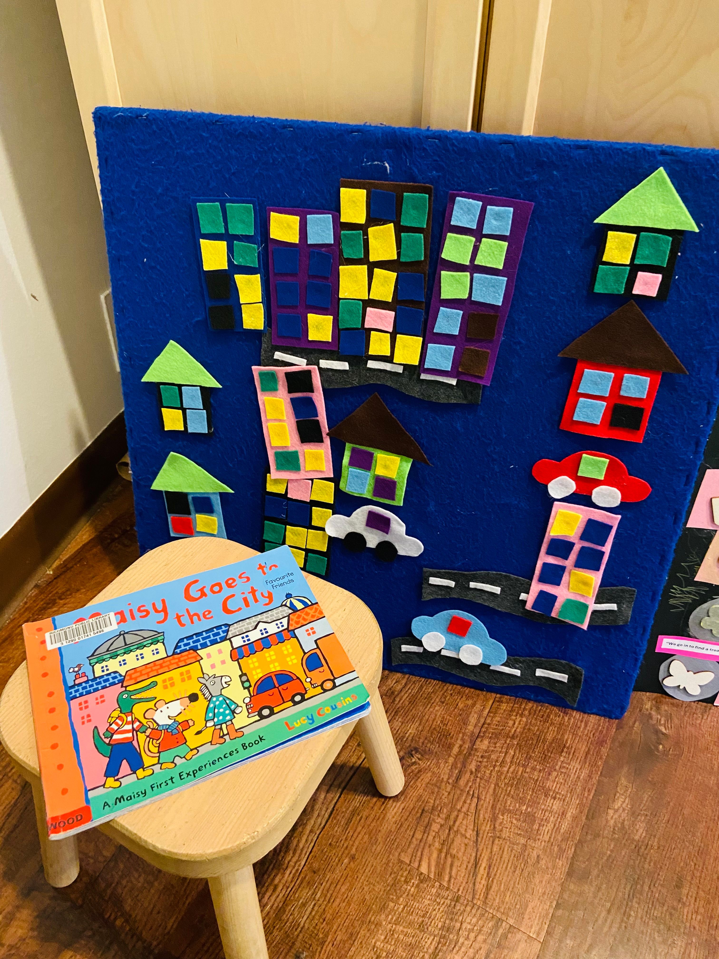 A felt city scene of houses, apartment blocks and cars on a blue board, with the picture book Maisy Goes to the City propped on a stool in front.