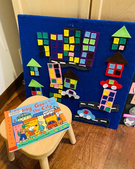 A felt city scene of houses, apartment blocks and cars on a blue board, with the picture book Maisy Goes to the City propped on a stool in front.