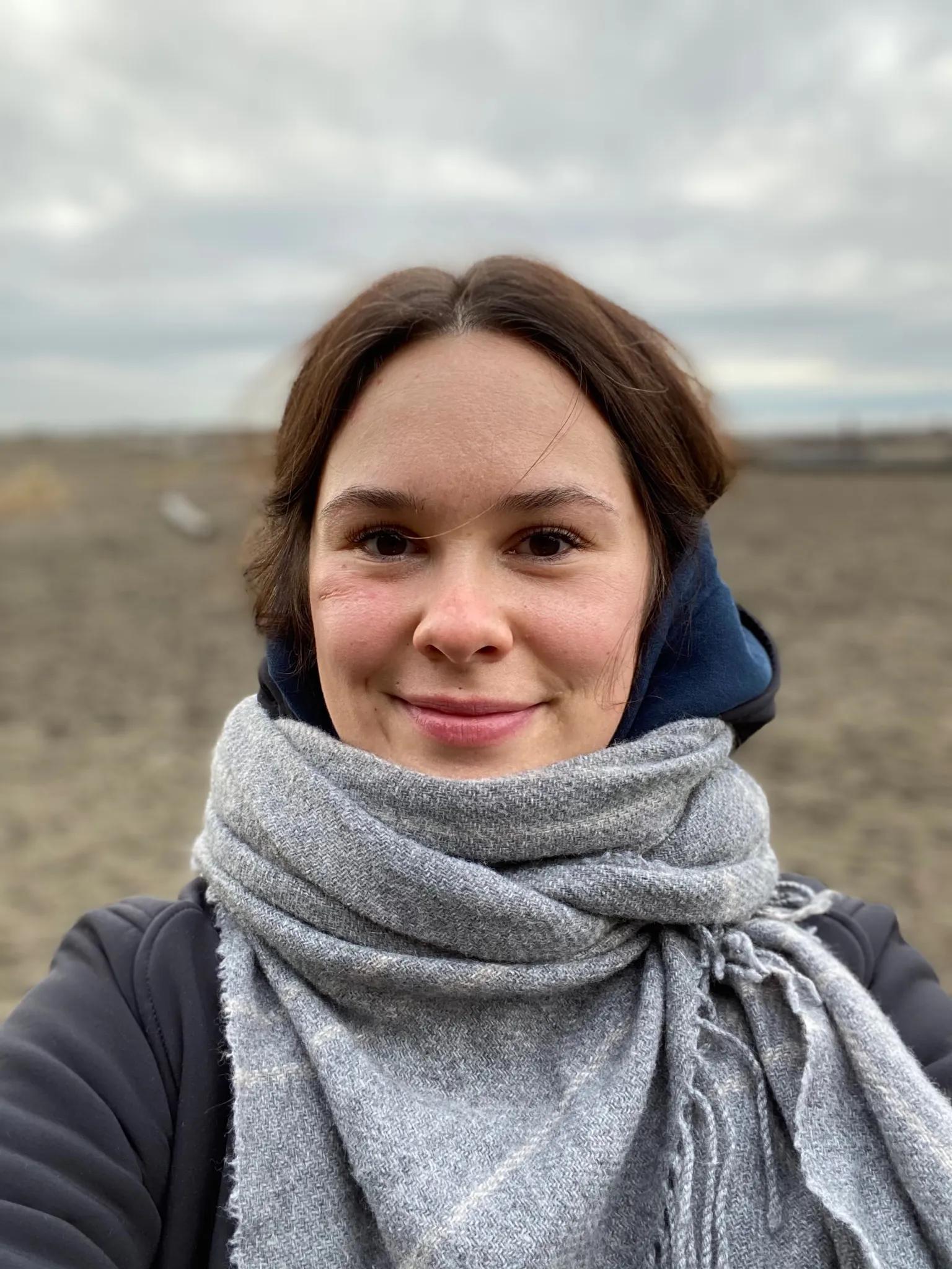 Cayley Burton outdoors on an overcast day, wrapped in a grey wool scarf and smiling at the camera.