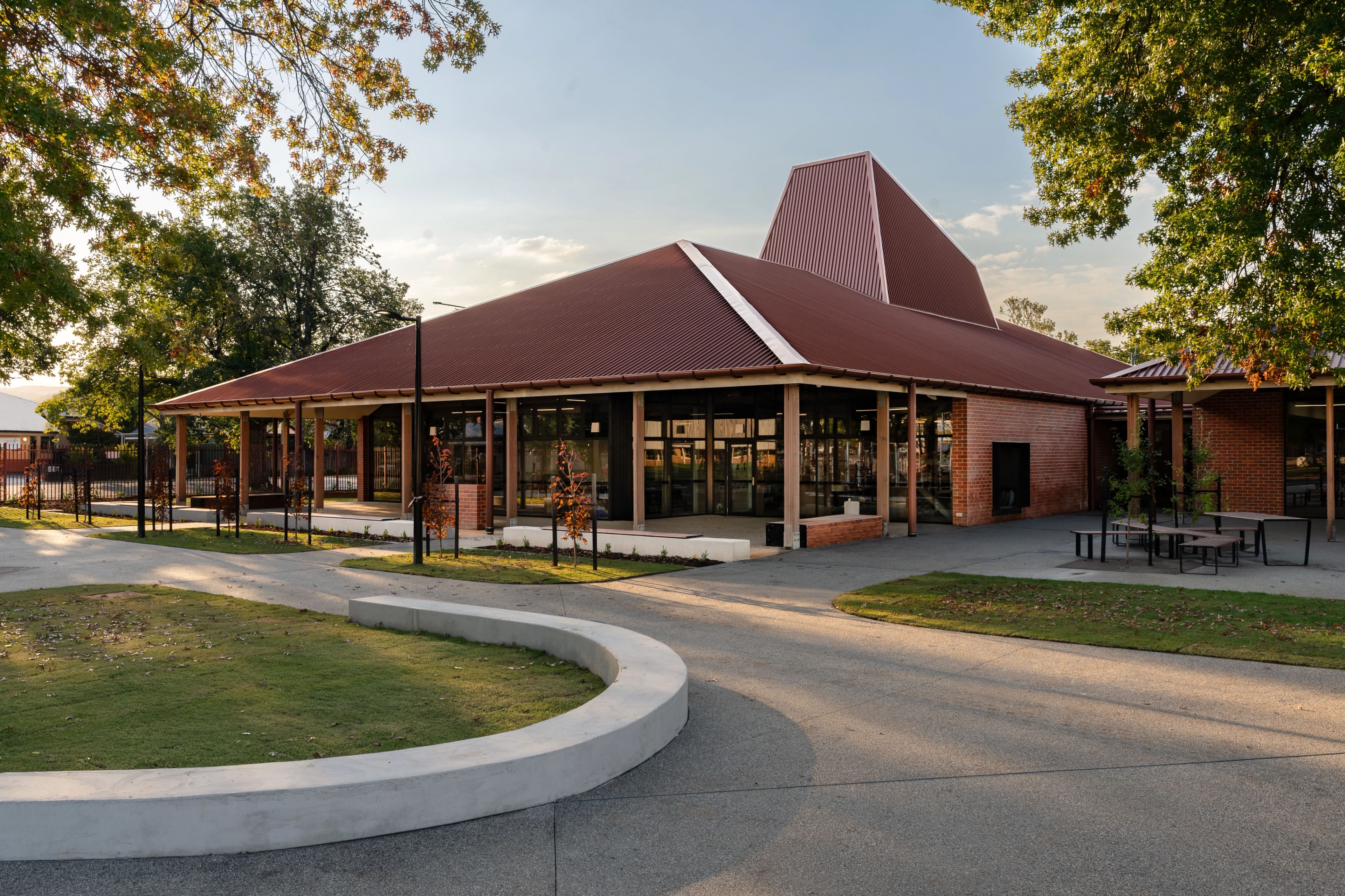 A view of The Scots School campus showcasing a contemporary school building with a distinctive red metal roof, exposed brickwork, timber columns and generous covered outdoor areas. The landscaped setting creates a welcoming connection between the building, outdoor spaces and school grounds.
