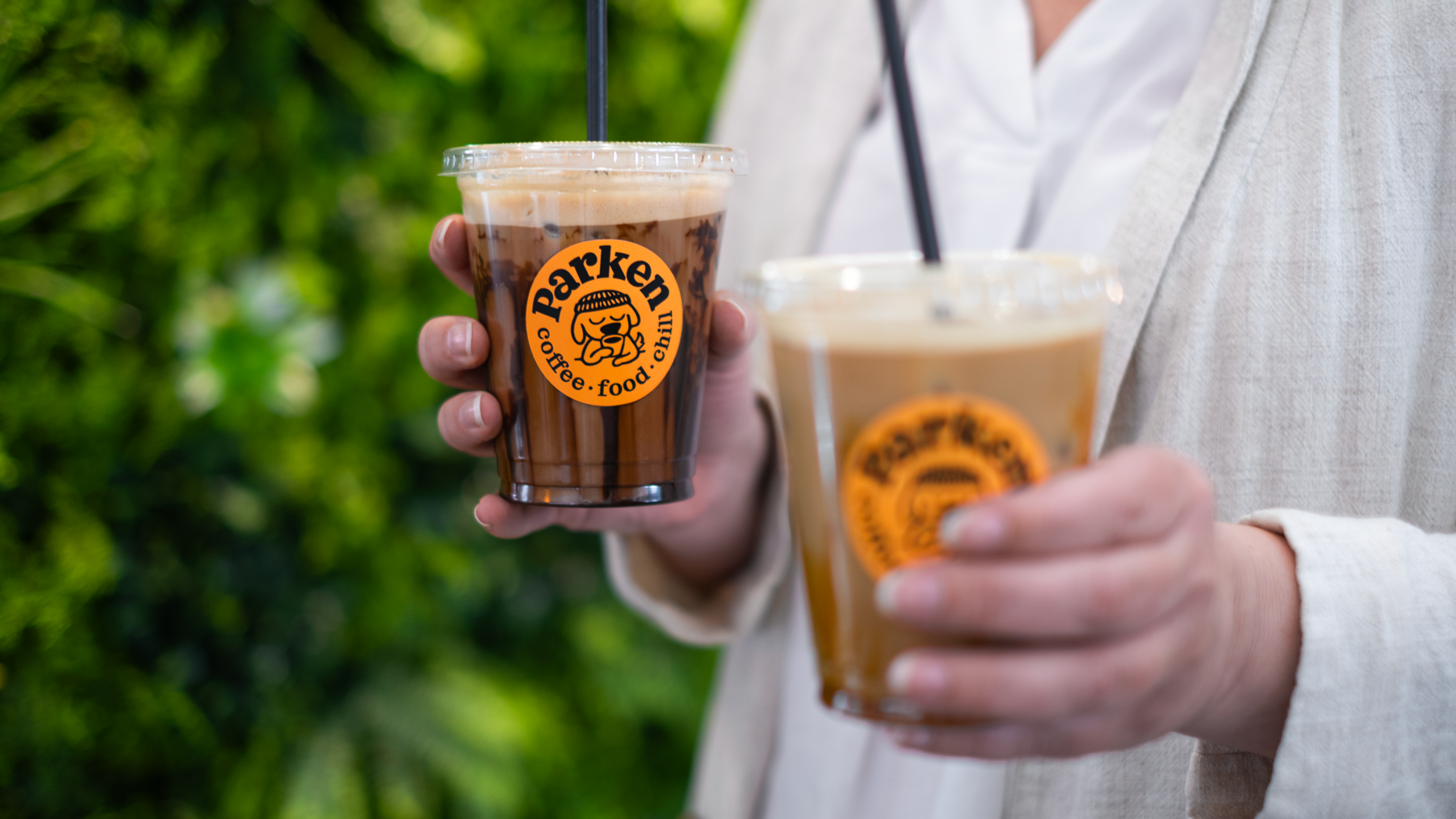 Parken Nærbilde av kvinne i lyse businessklær som holder to iskaffe med oransje Parken logo sticker, med grønn bakgrunn. En Islatte og en iskaffe mocka.