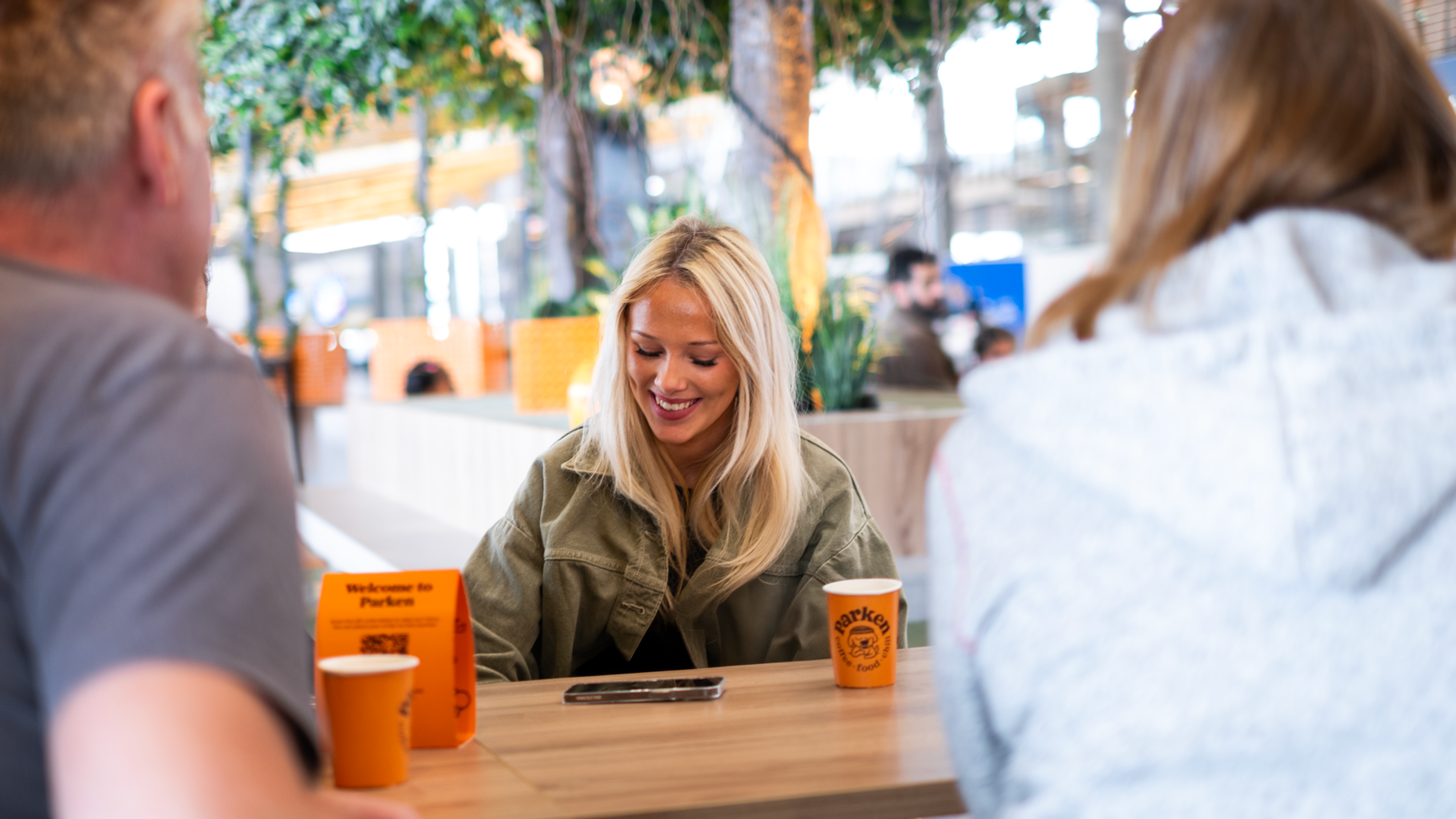 Parken stemingsfoto av ung blond jente som sitter på en koselig cafe på gardemoen, hun smiler og sitter ved et bord med Parken kaffekopper. Vi ser ryggen til to mennesker i blurr i forgrunn. I bakgrunn ser vi Parken. medhyggelig og grønne omgivelser. 