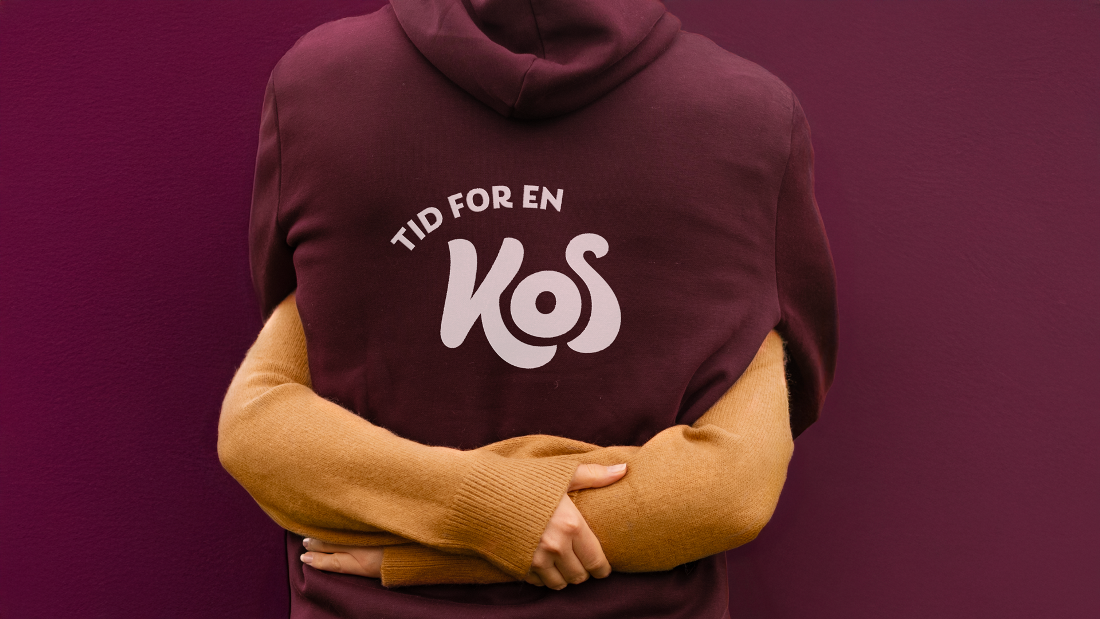 Bildet viser to som klemmer hverandre. Hen ene har genser med kos logo