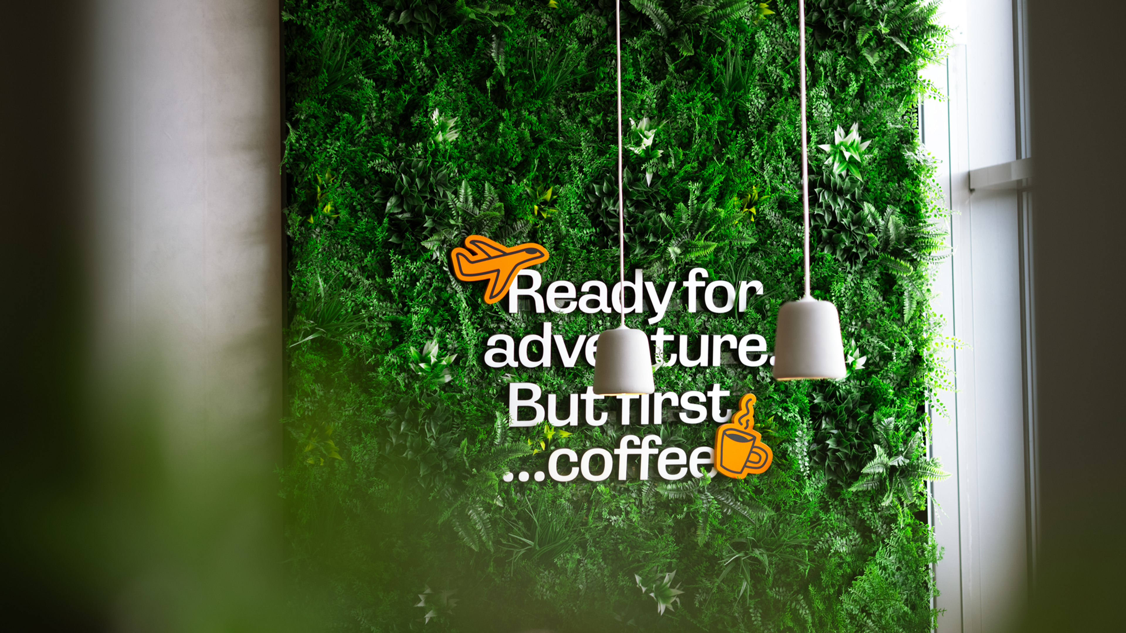 Parken grønn plantevegg med hvit tekst av quote "Ready for adventure. But first coffee", plassert oppå håndtegnet fly ikon og kaffe ikon i orange som matcher Parken grafiske profil. 