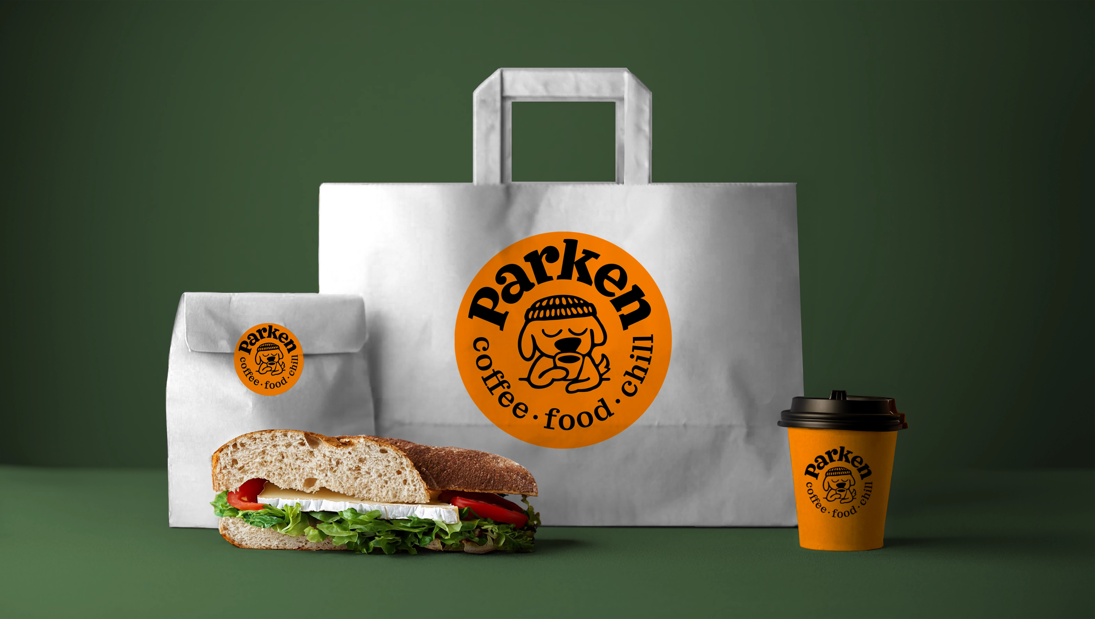 Parken Takeaway pakning på grønn bakgrunn. Hvit pose med oransje Parken logo, liten og lukket takeaway pose med oranje sticker med Parken logo. Tiø høyre oransje kaffekopp med Parken logo. I front en sandwich med salat og brie. 