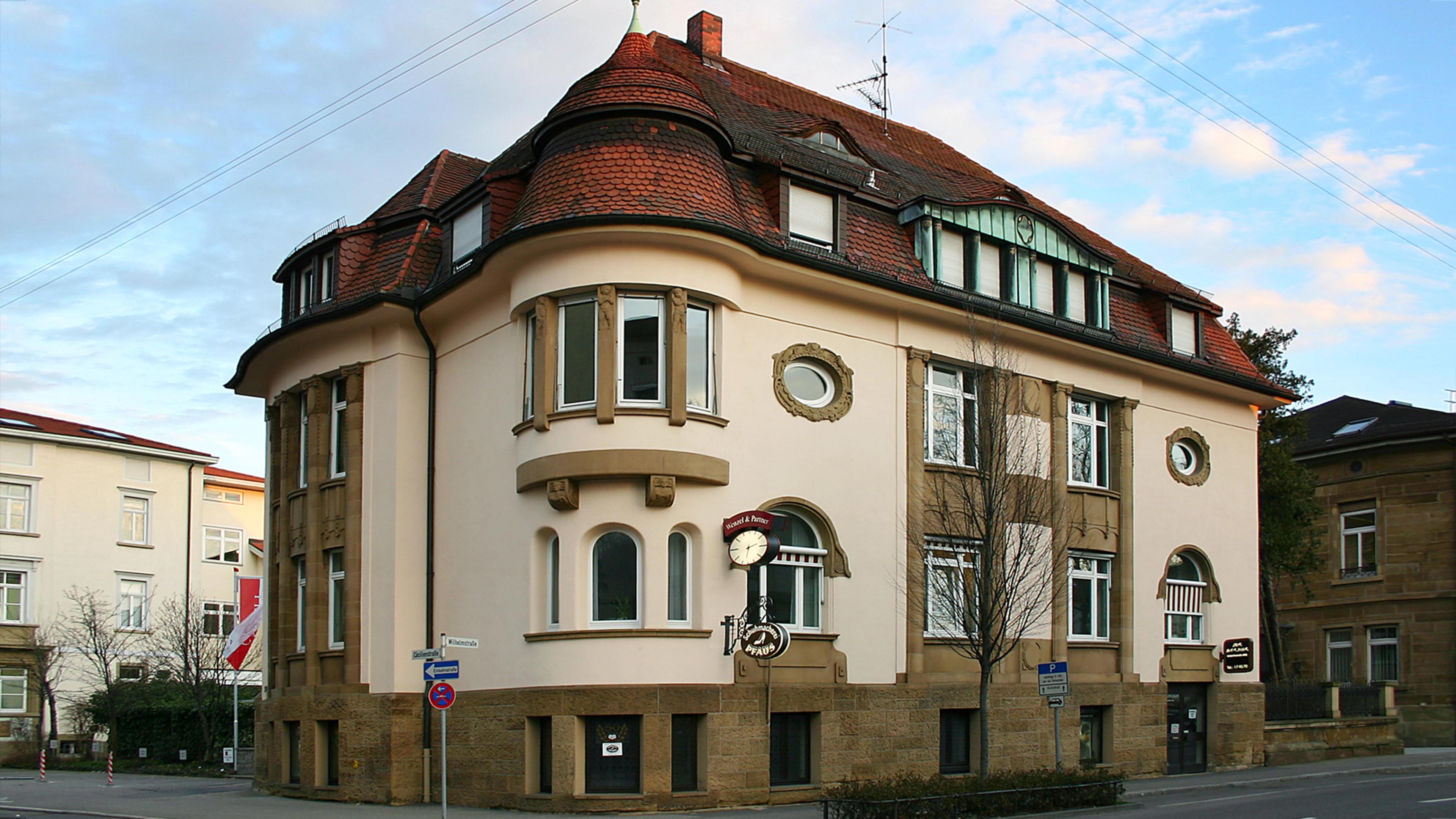 Historisches Gebäude mit abgerundeter Ecke, roten Dachziegeln und mehreren Fenstern in einer städtischen Umgebung