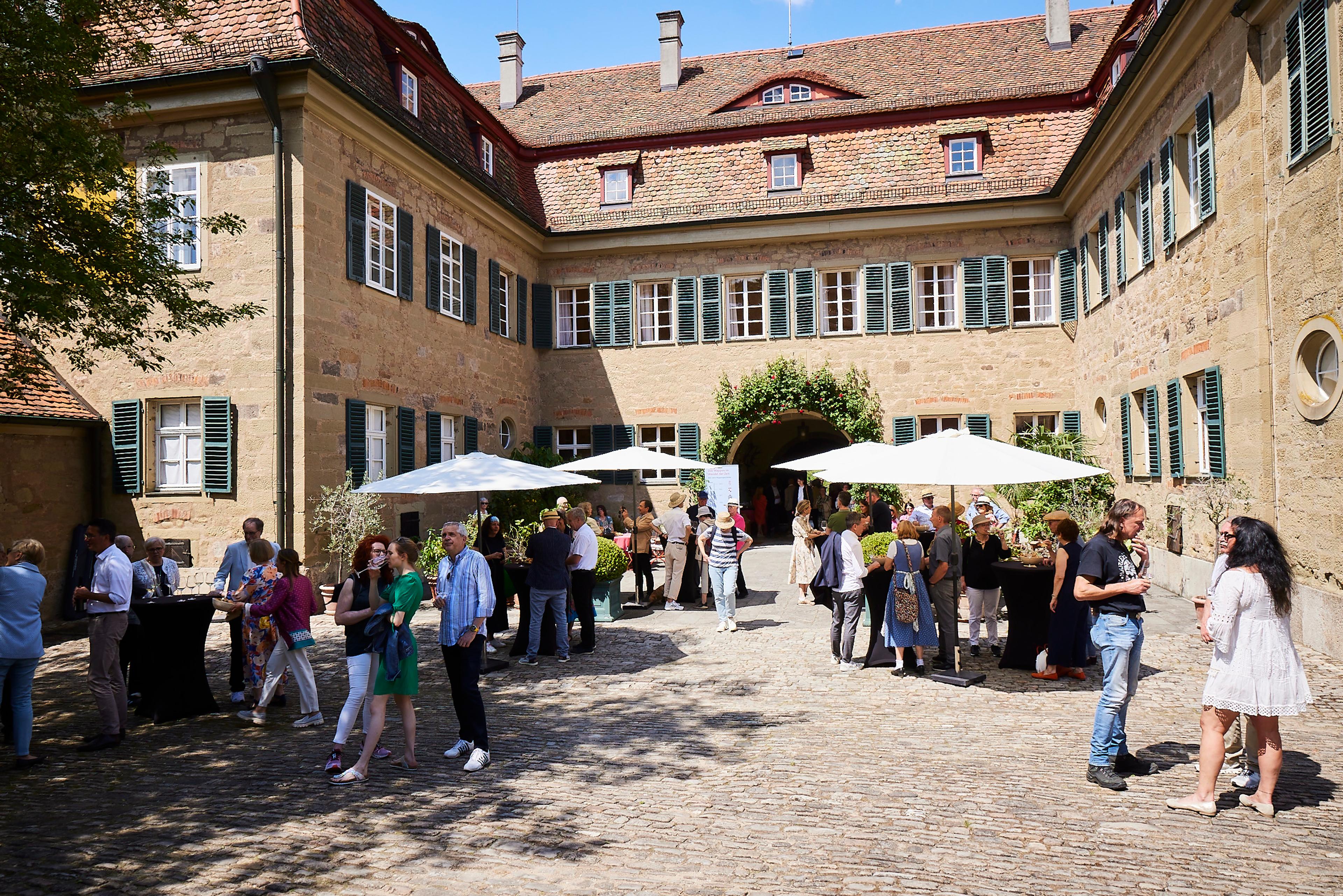 Menschen unterhalten sich und stehen an Stehtischen mit weißen Sonnenschirmen im Innenhof eines historischen Gebäudes mit Sandsteinmauern und grünen Fensterläden