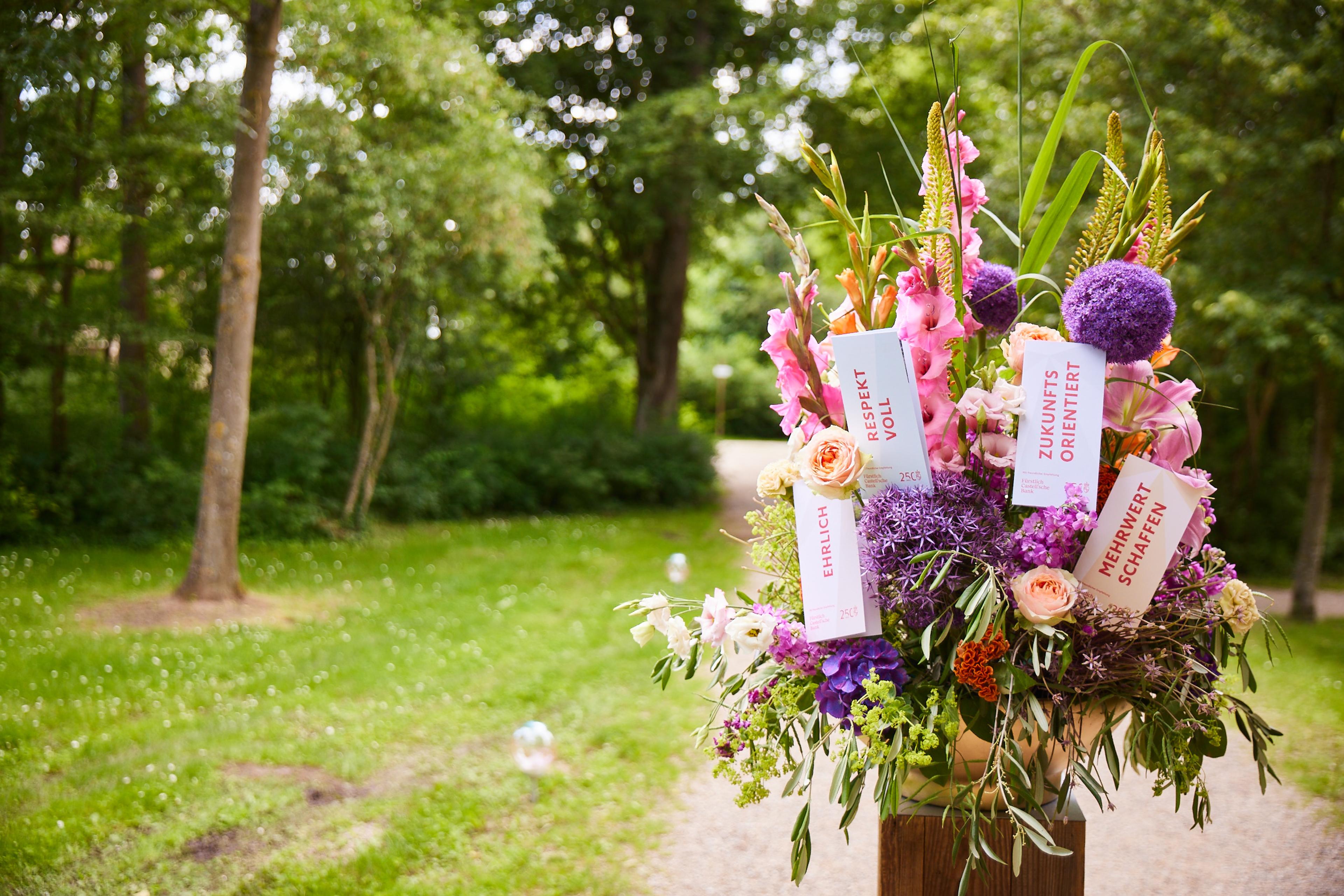 Bunter Blumenstrauß mit rosa, lila und weißen Blumen im Park, beschriftete Karten mit Worten wie 'Zukunftsorientiert' und 'Mehrwert schaffen', 'Respektvoll' und 'Ehrlich'