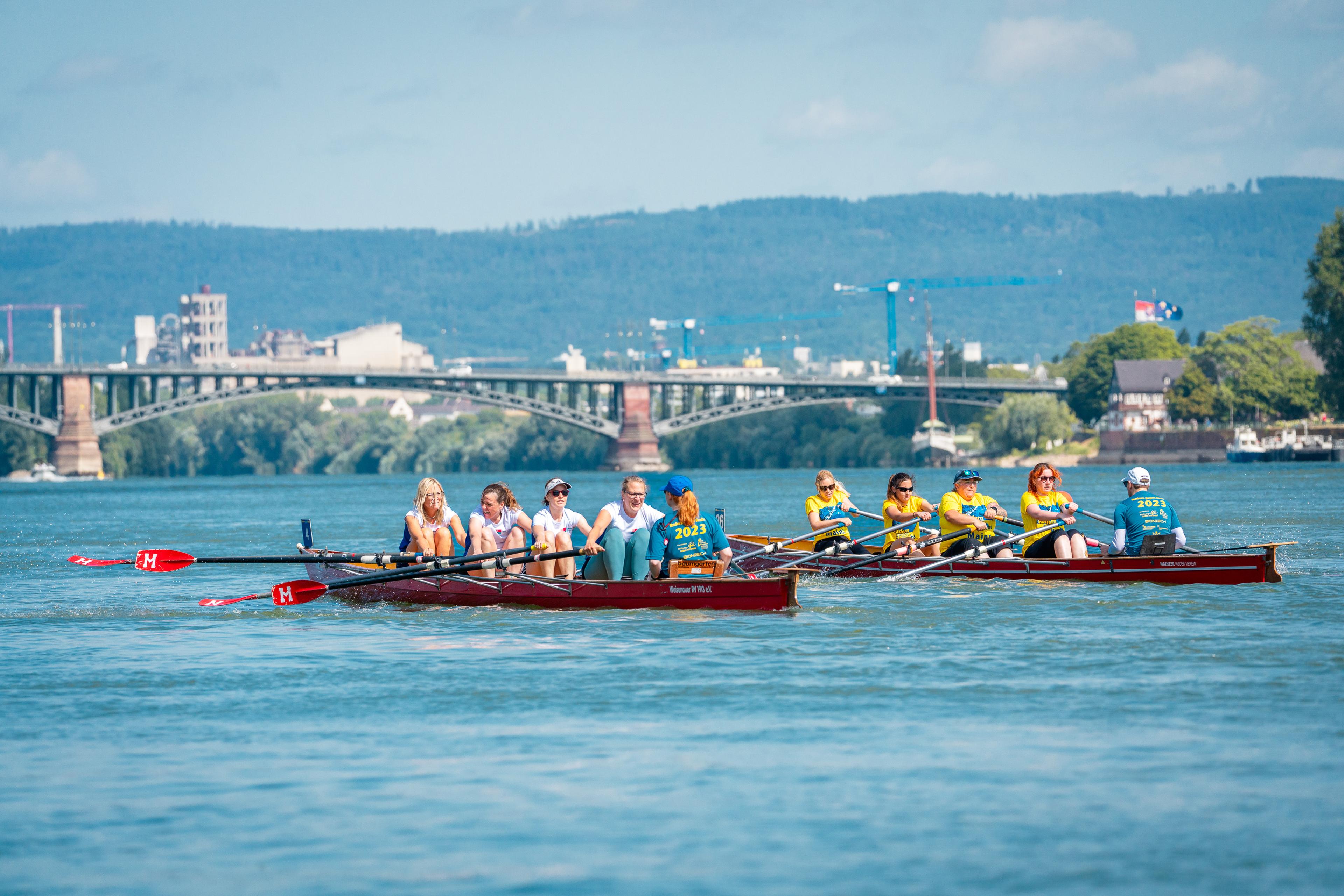 Zwei Ruderboote mit jeweils acht Ruderern auf einem Fluss, im Hintergrund eine Brücke und bewaldete Hügel