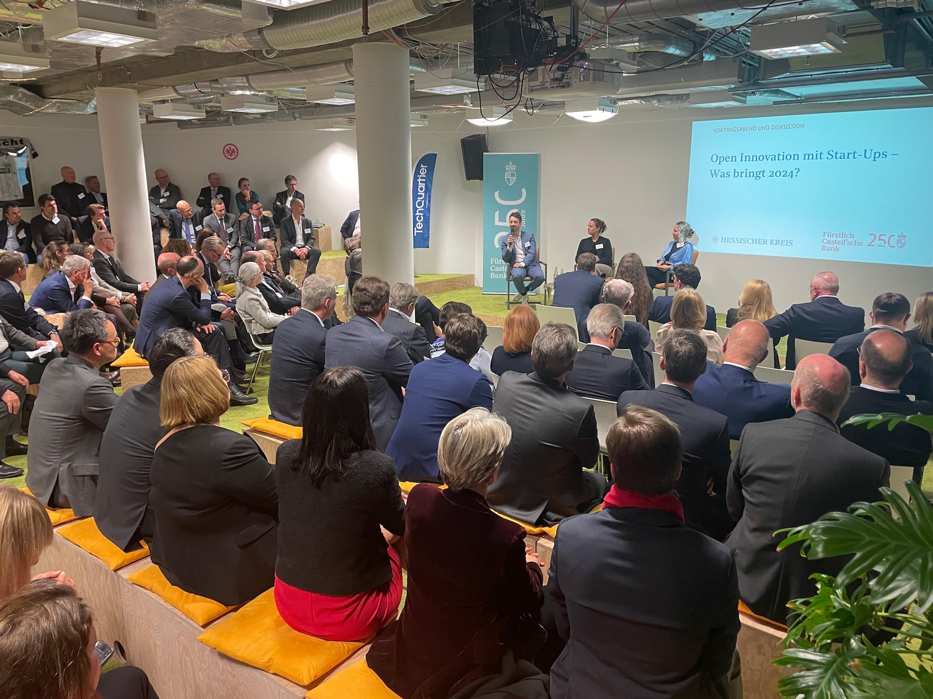 Große Gruppe von Menschen verfolgt Podiumsdiskussion zum Thema 'Open Innovation mit Start-Ups – Wie bringt man es?' in modernem Veranstaltungsraum