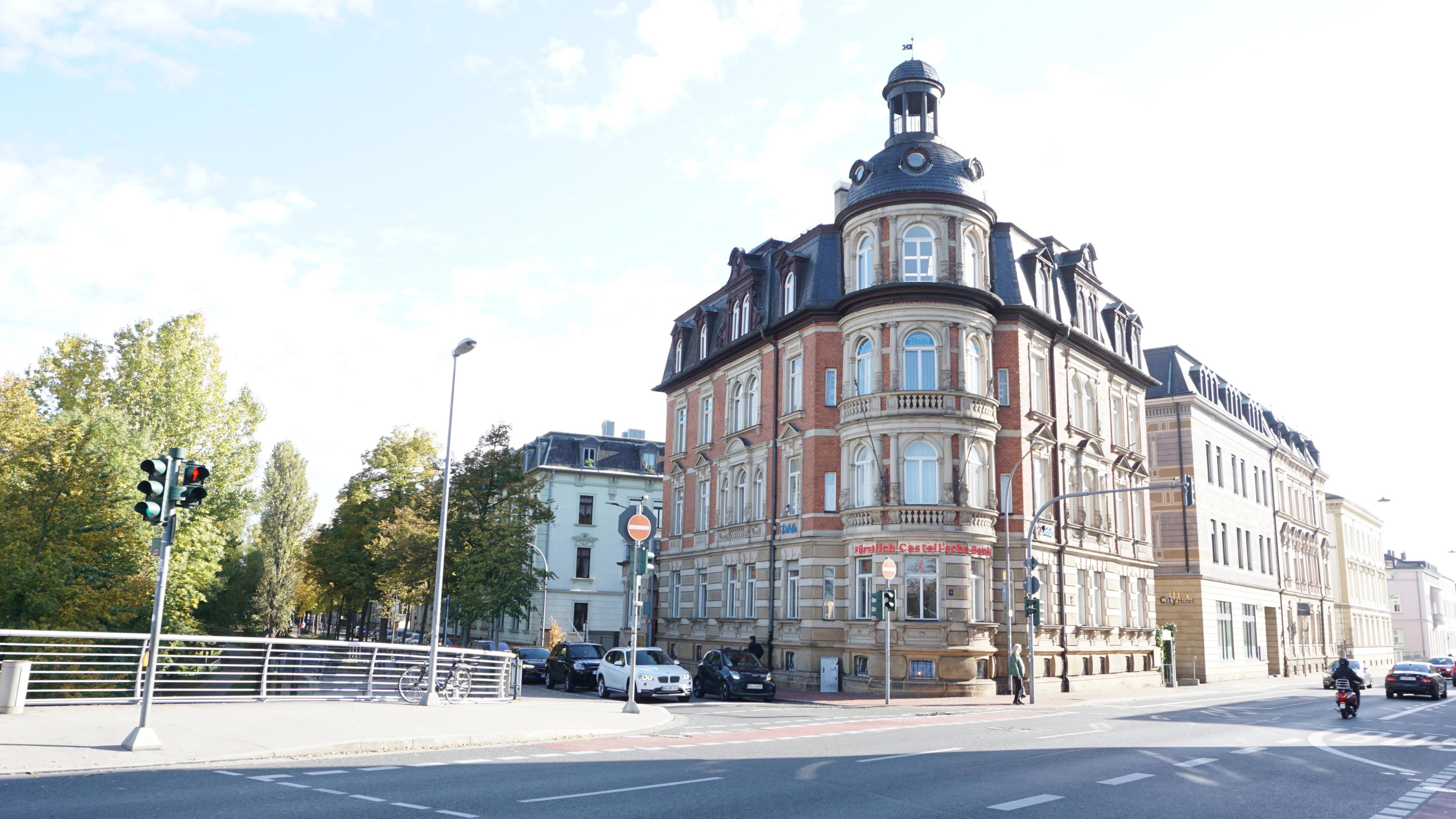 Historisches Eckgebäude mit Turm und Kuppel an einer Kreuzung, umgeben von Bäumen und Straßenverkehr