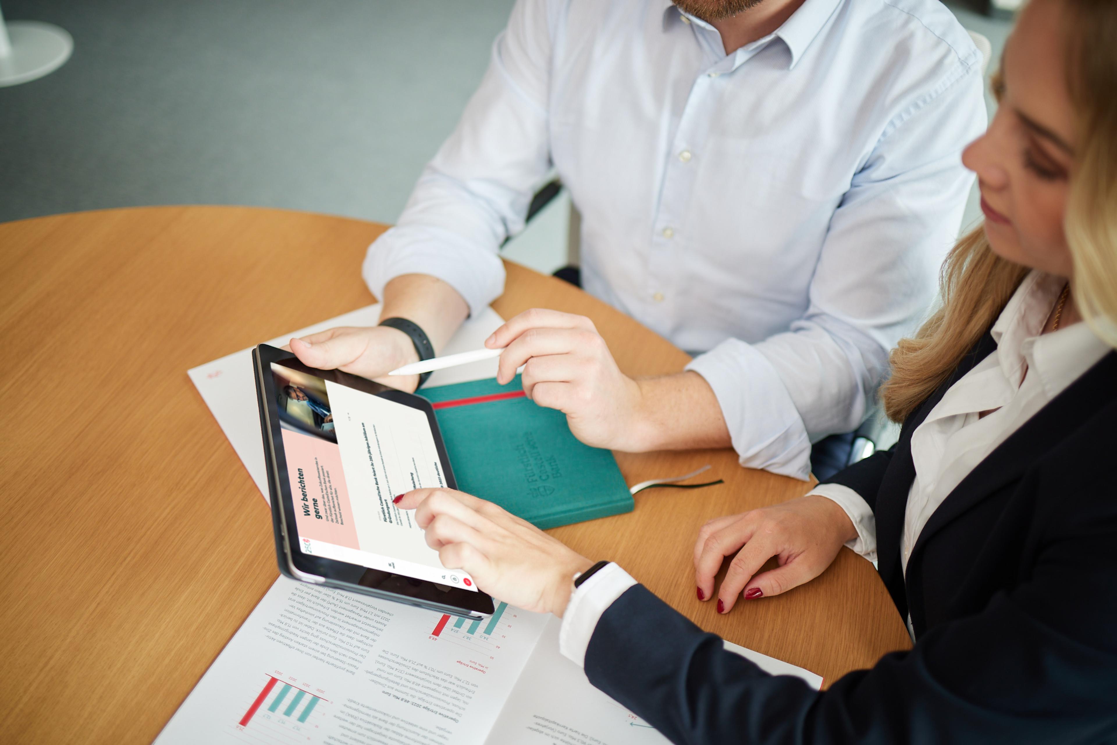 Zwei Personen besprechen Diagramme und Daten auf einem Tablet und ausgedruckten Dokumenten an einem Holztisch