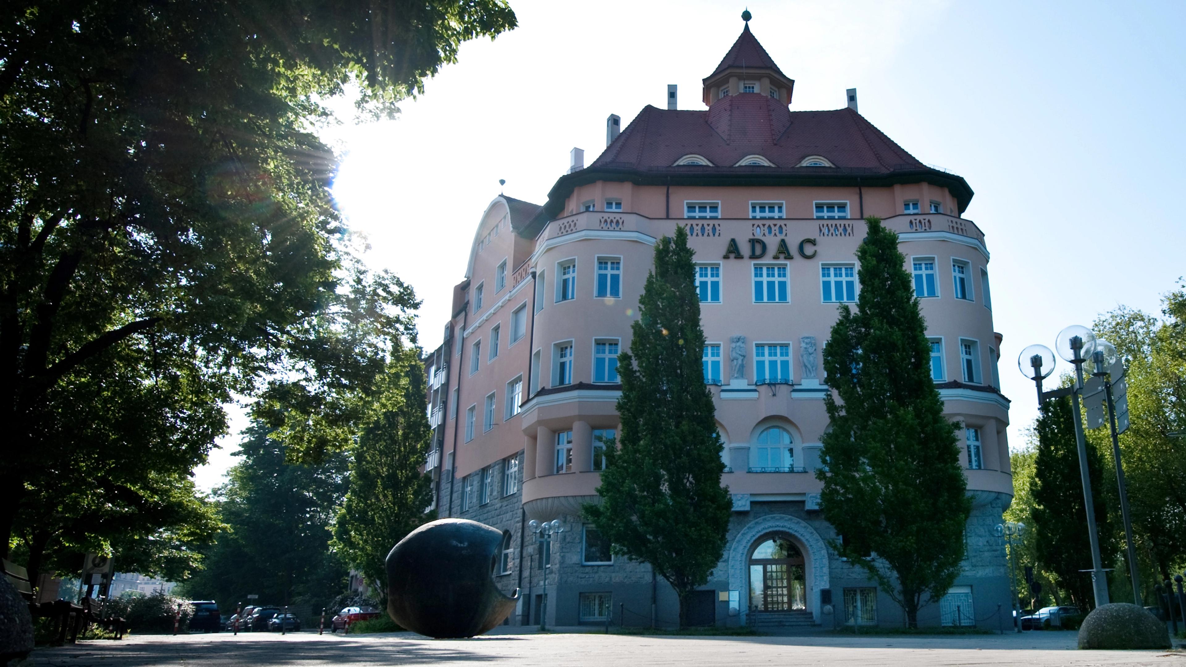 Historisches Gebäude mit rotem Dach und drei hohen Bäumen im Vordergrund, umgeben von grünen Bäumen bei Sonnenschein