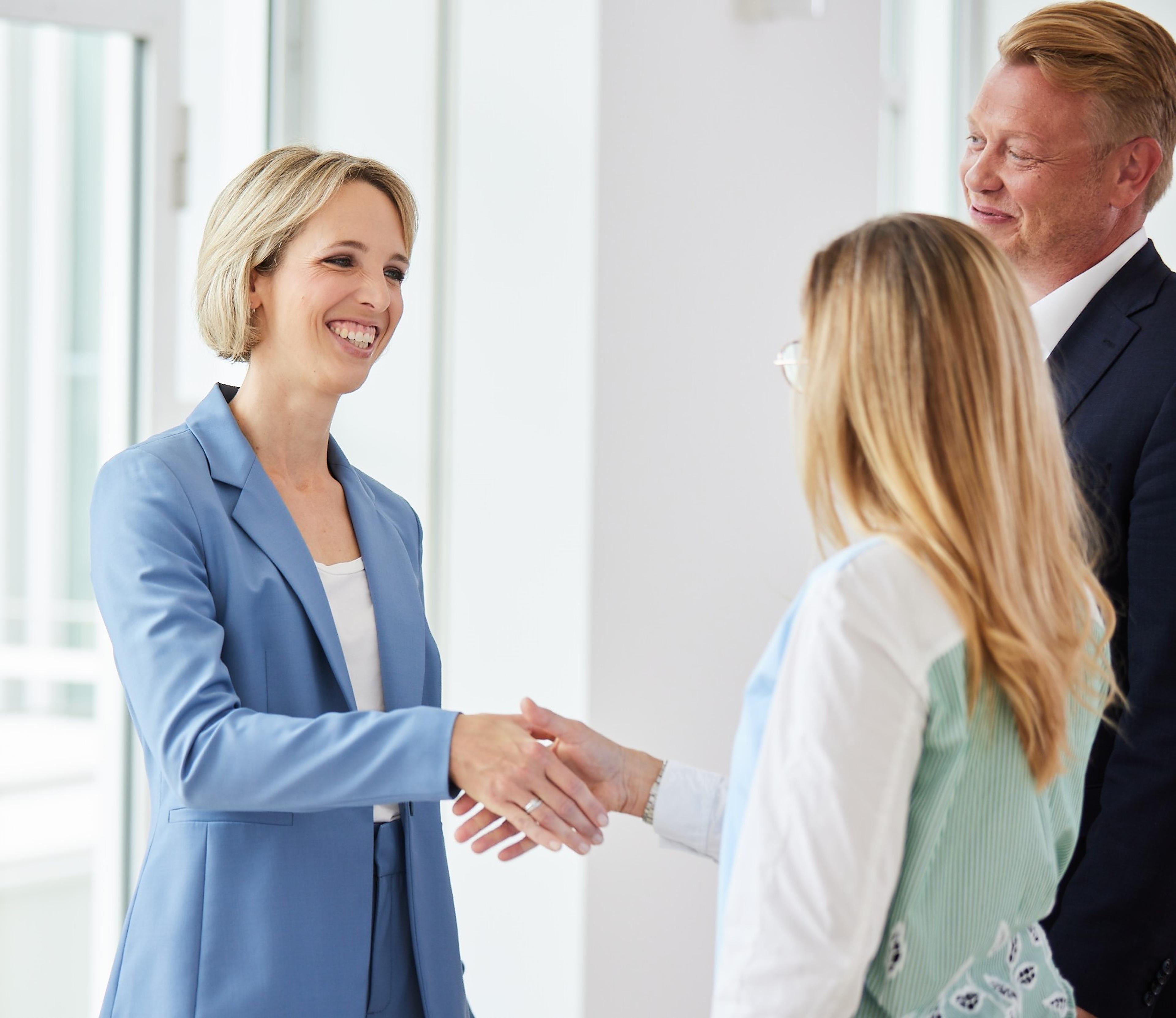 Zwei Frauen geben sich die Hand in einem hellen Büro, ein Mann lächelnd im Hintergrund