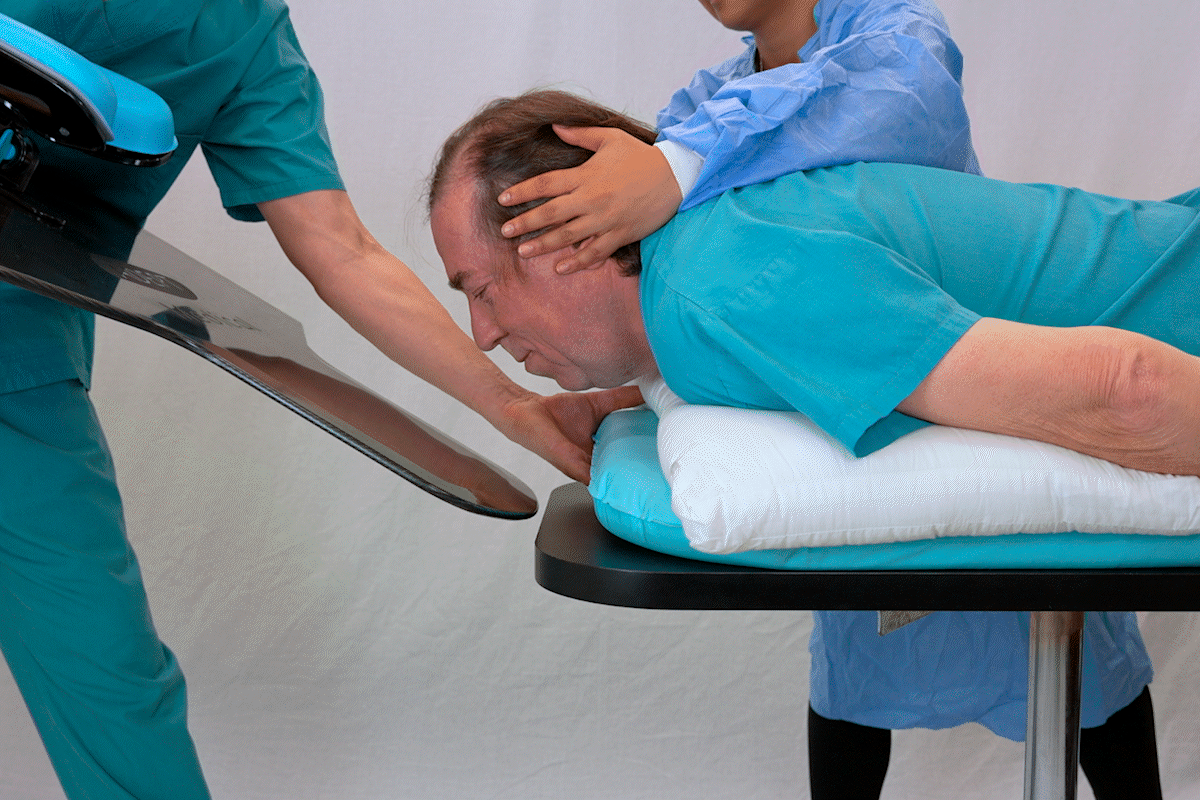 Man lying prone on a medical table, assisted by two medical staff, one holding a reflective board, the other supporting his head on a cushion.