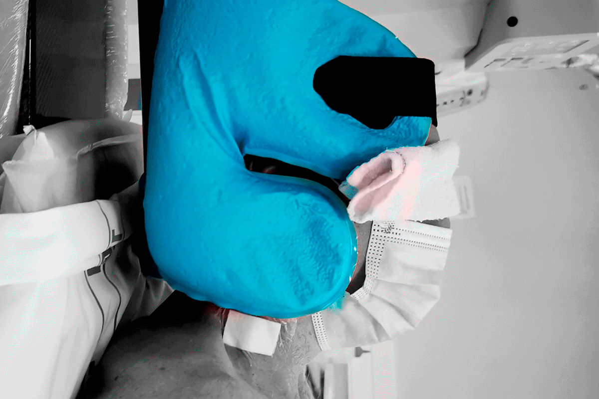 A person wearing a surgical mask and a bright blue medical head device, shown in color against a grayscale hospital background.