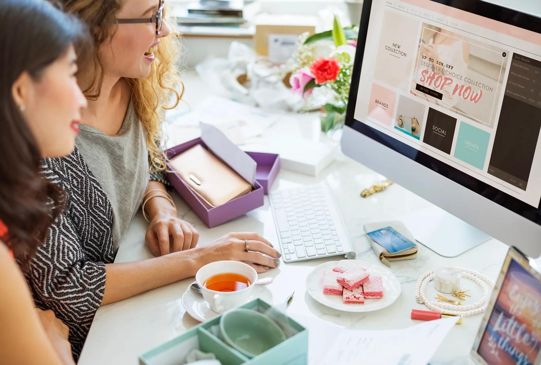 Two women sitting side by side at a desk and looking at an online shop on the screen