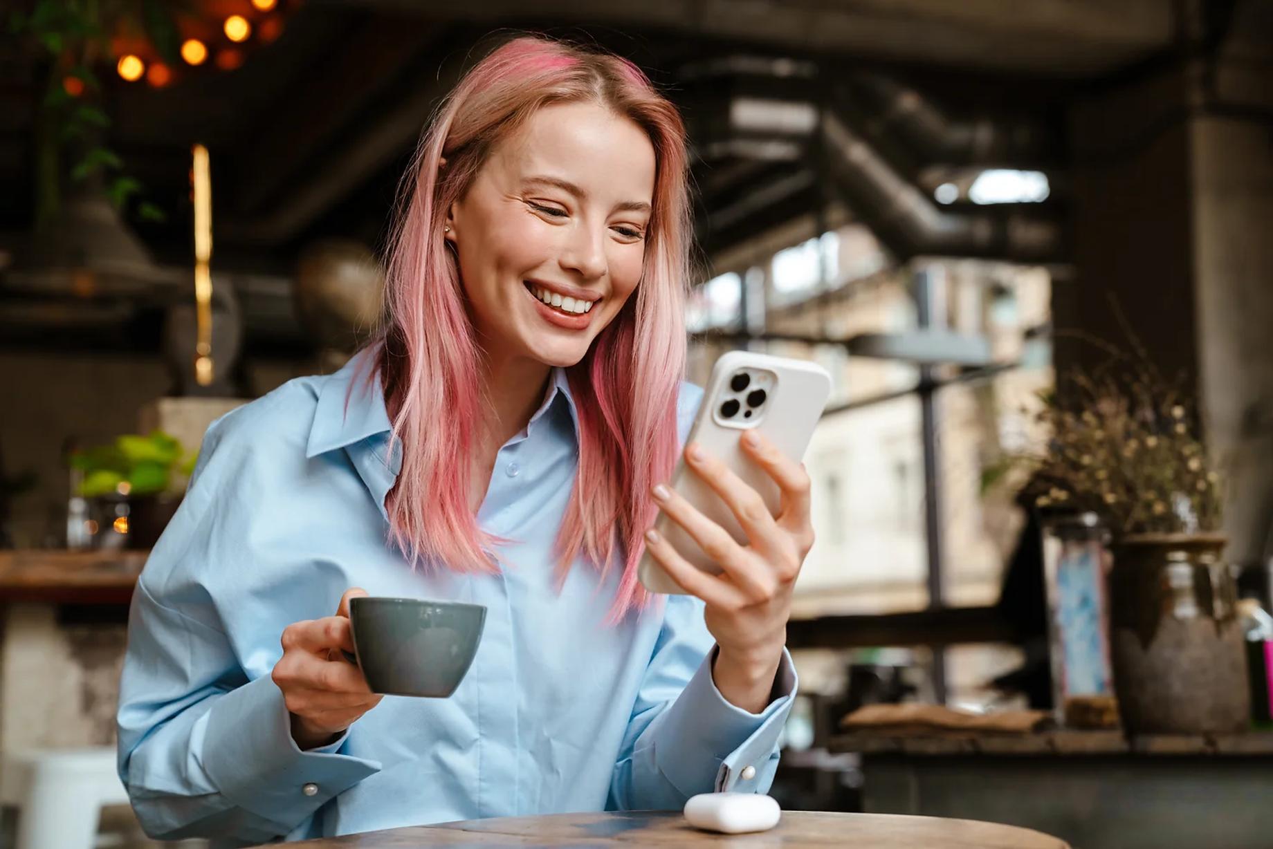 A lady with pink hair sitting in a coffee shop looking at a white smartphone in her hand