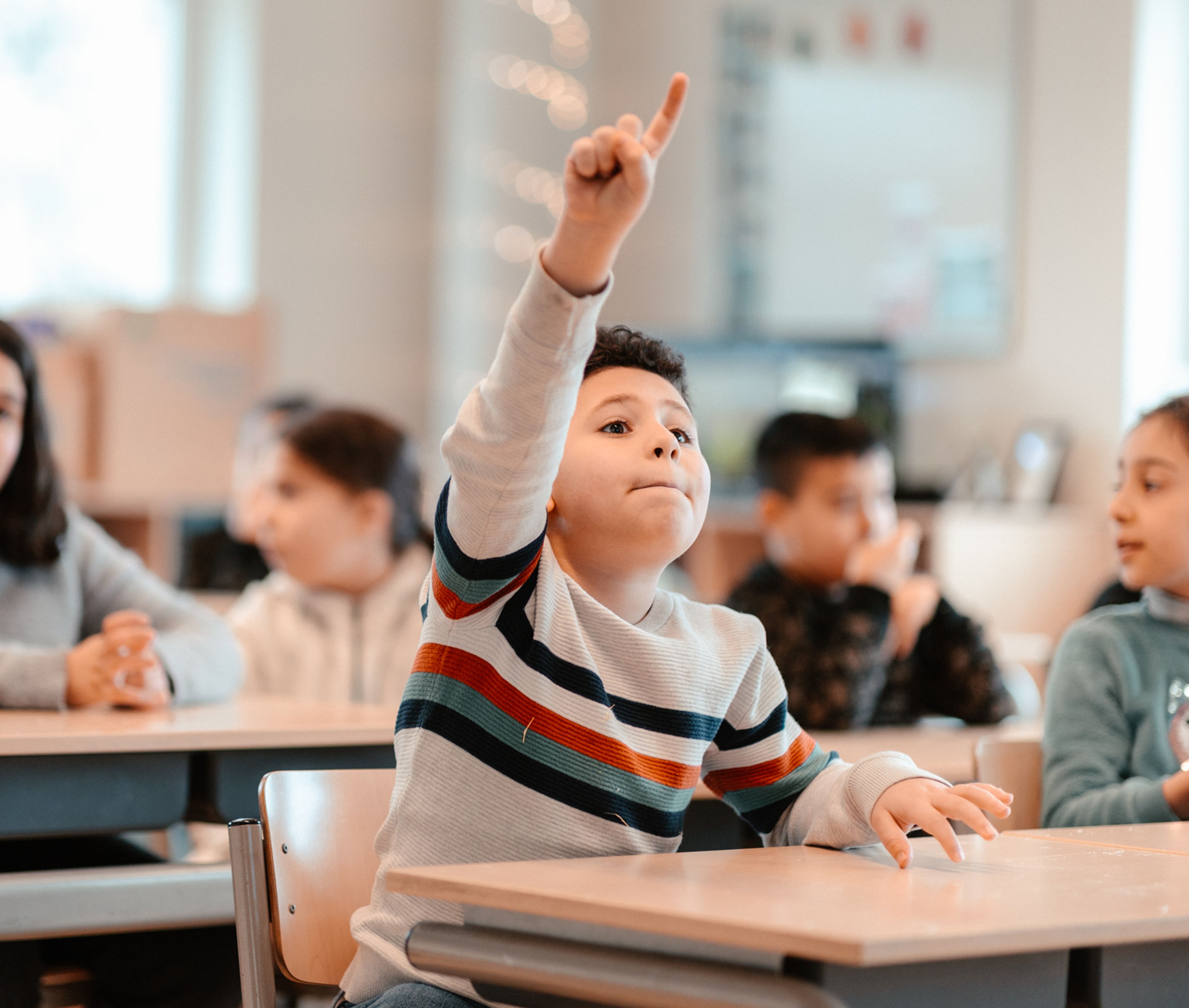 Foto van klaslokaal met kinderen die aan tafels zitten, waarbij een kind met gestreepte trui zijn hand omhoog steekt. Context toont actieve deelname en interactie tijdens lesmoment.