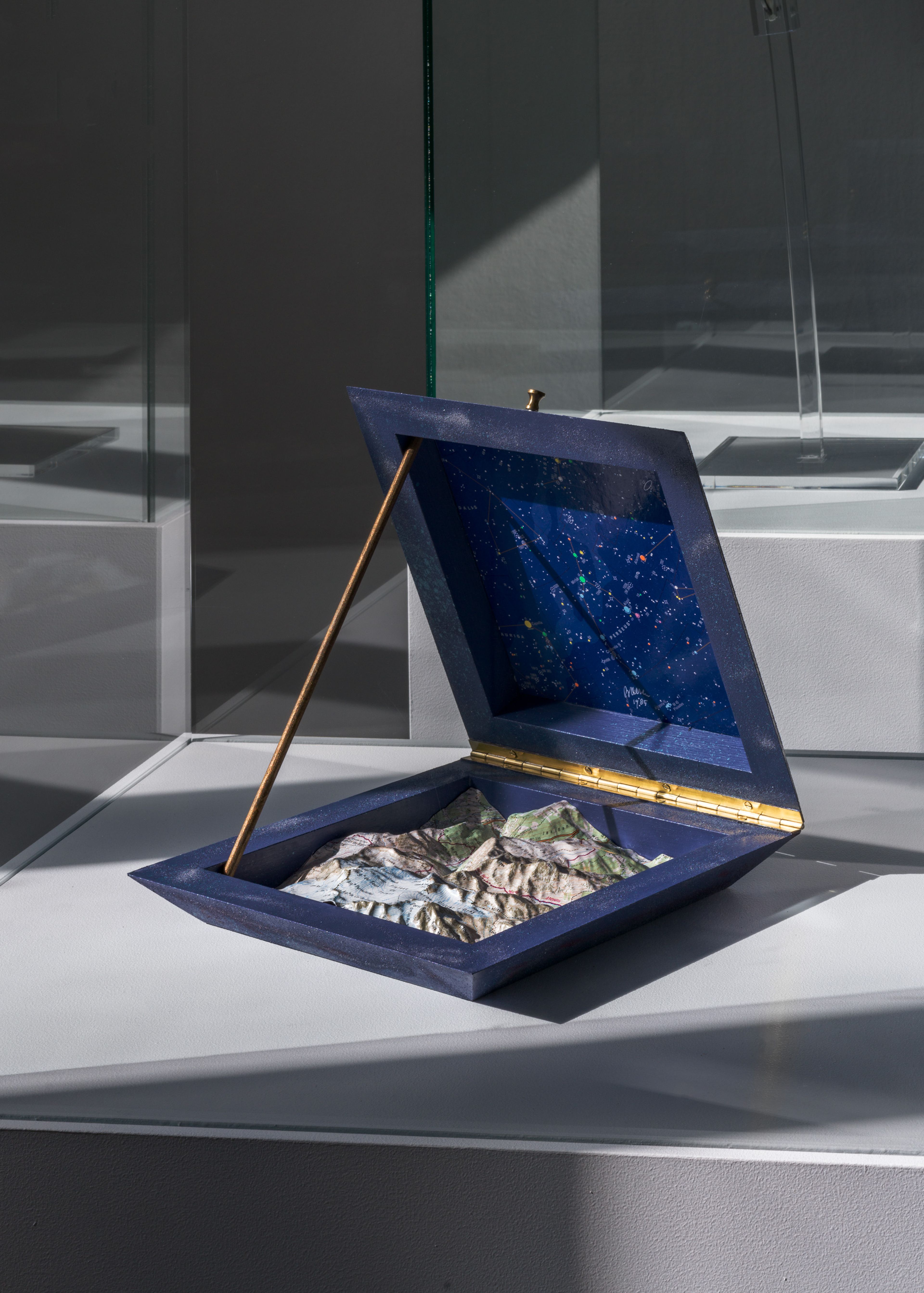 Marinus Boezem, _Della Scultura & La Luce_, 1985 Foto van een geopende driedimensionale hemel- en landkaart in een blauw vierkant doosje met gouden scharnieren en steun. Bovenkant toont sterrenbeeldkaart met sterren en lijnen, onderkant reliëfkaart met bergen en landschap.