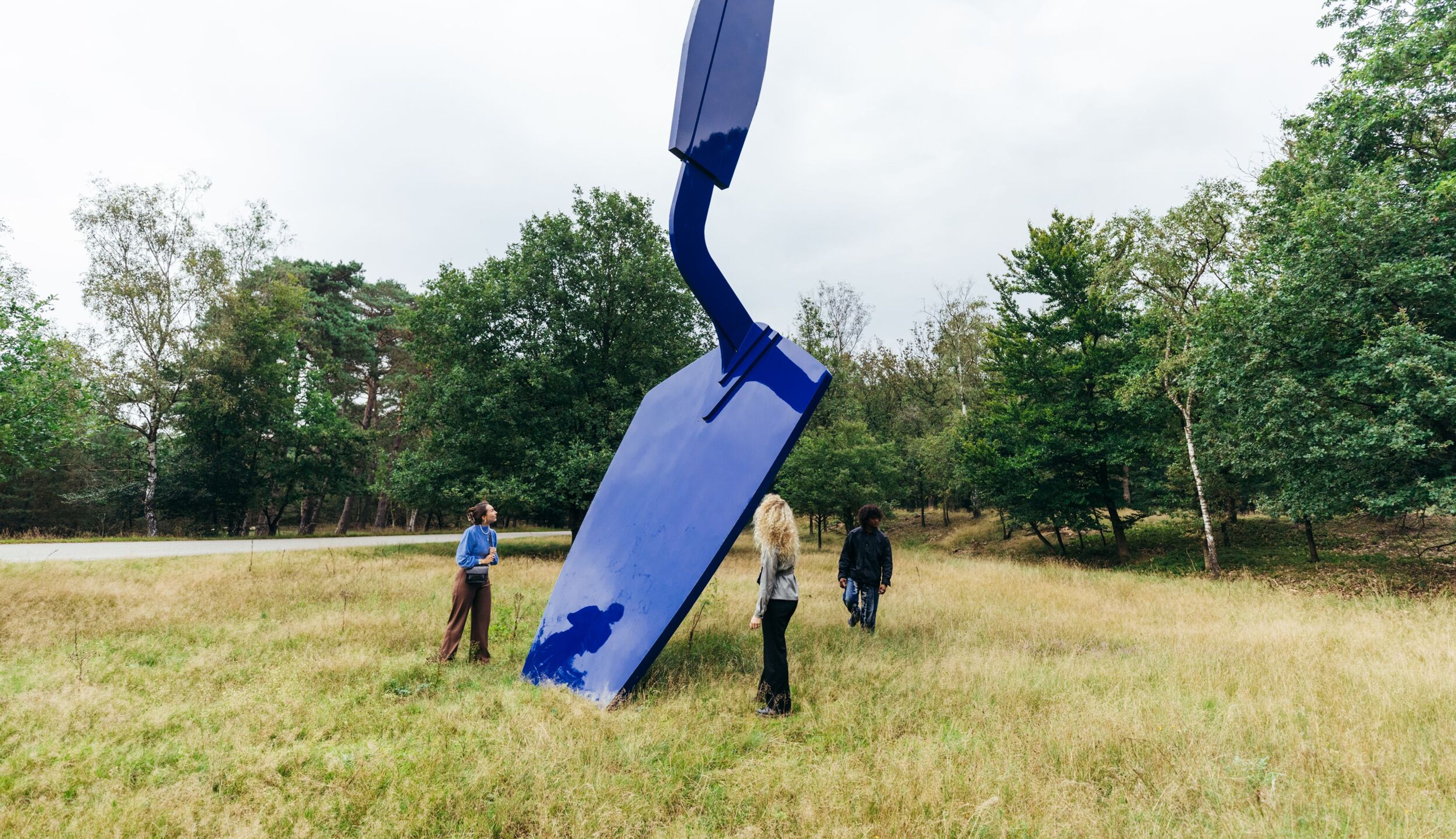 Foto van een grote blauwe sculptuur geplaatst in een grasveld omringd door bomen, met drie mensen die het kunstwerk bekijken.
