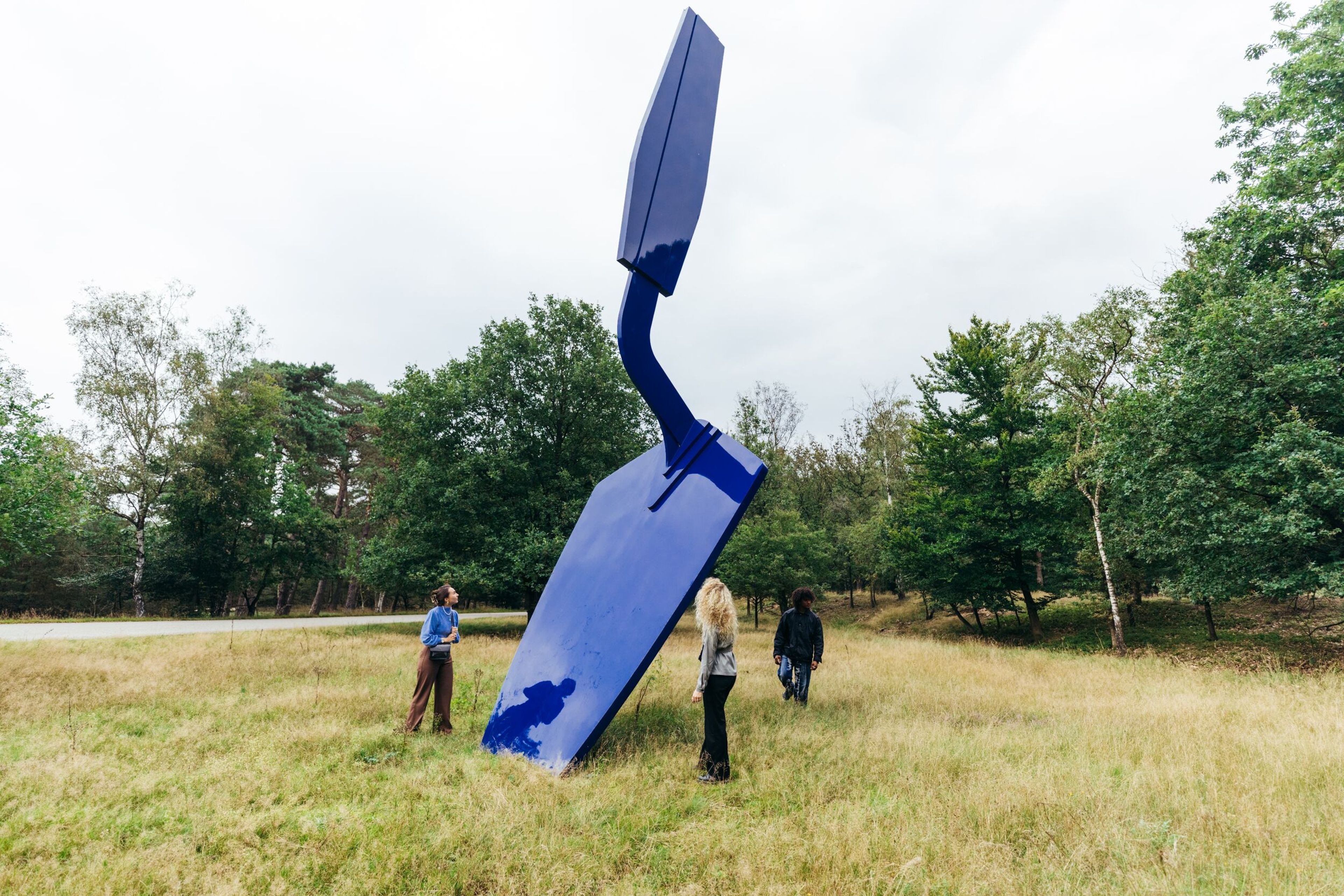 Foto van een grote blauwe sculptuur geplaatst in een grasveld omringd door bomen, met drie mensen die het kunstwerk bekijken.