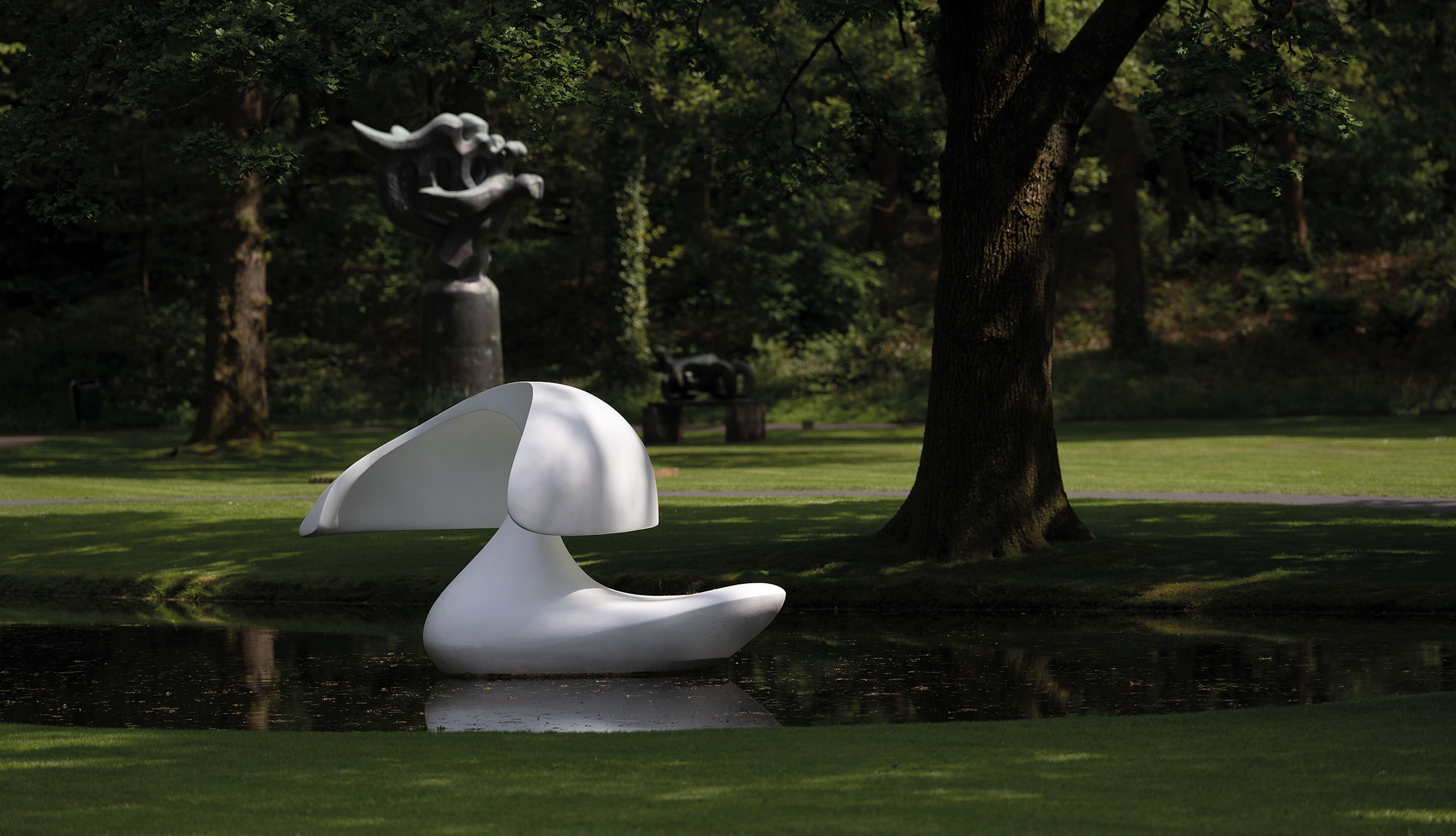 Foto van een moderne, witte abstracte sculptuur geplaatst op een wateroppervlak in een parkachtige omgeving met gras en bomen. Op de achtergrond staat nog een sculptuur.