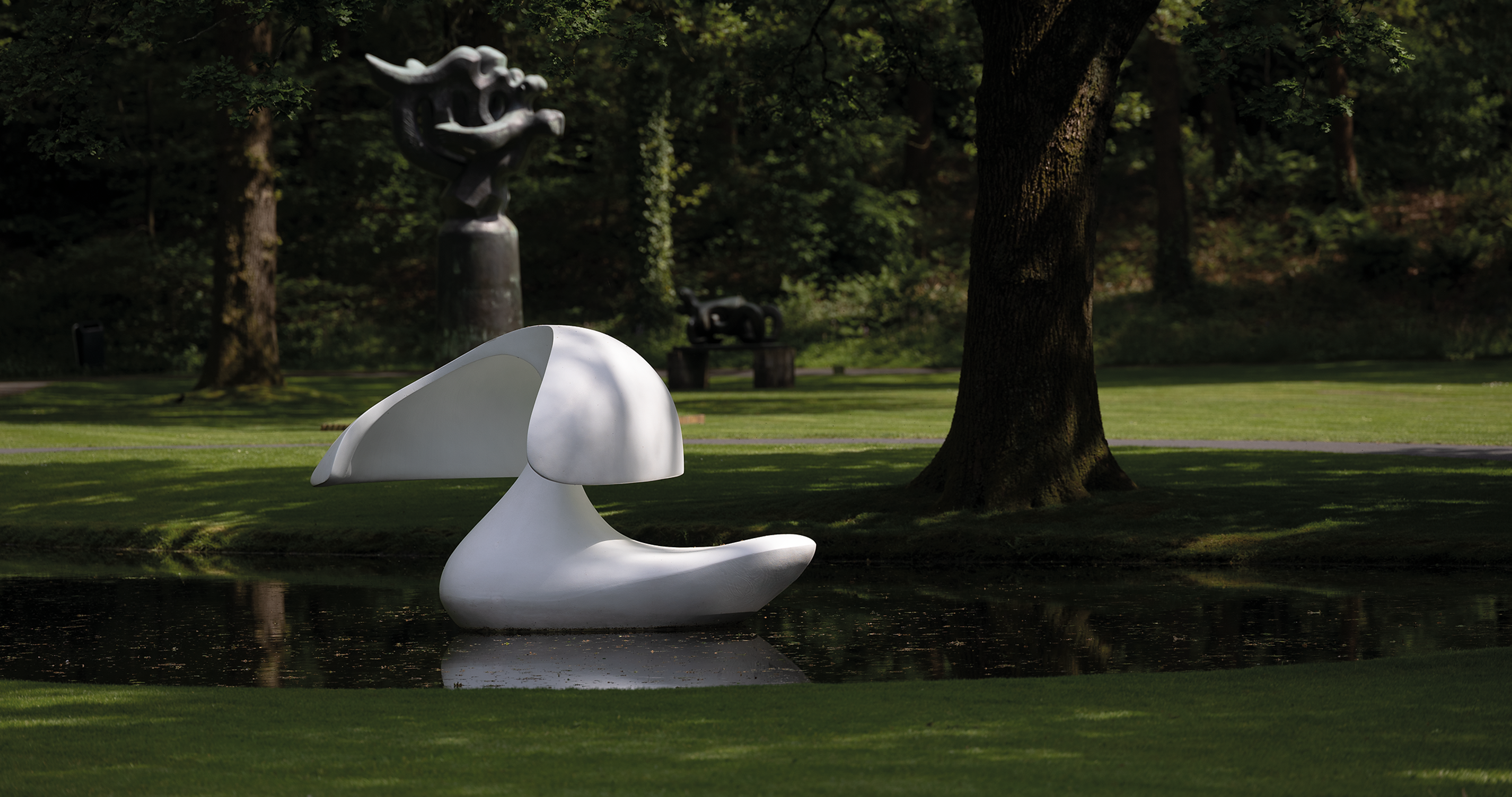 Marta Pan, _Sculpture flottante, Otterlo_, 1960-1961 Foto van een moderne, witte abstracte sculptuur geplaatst op een wateroppervlak in een parkachtige omgeving met gras en bomen. Op de achtergrond staat nog een sculptuur.