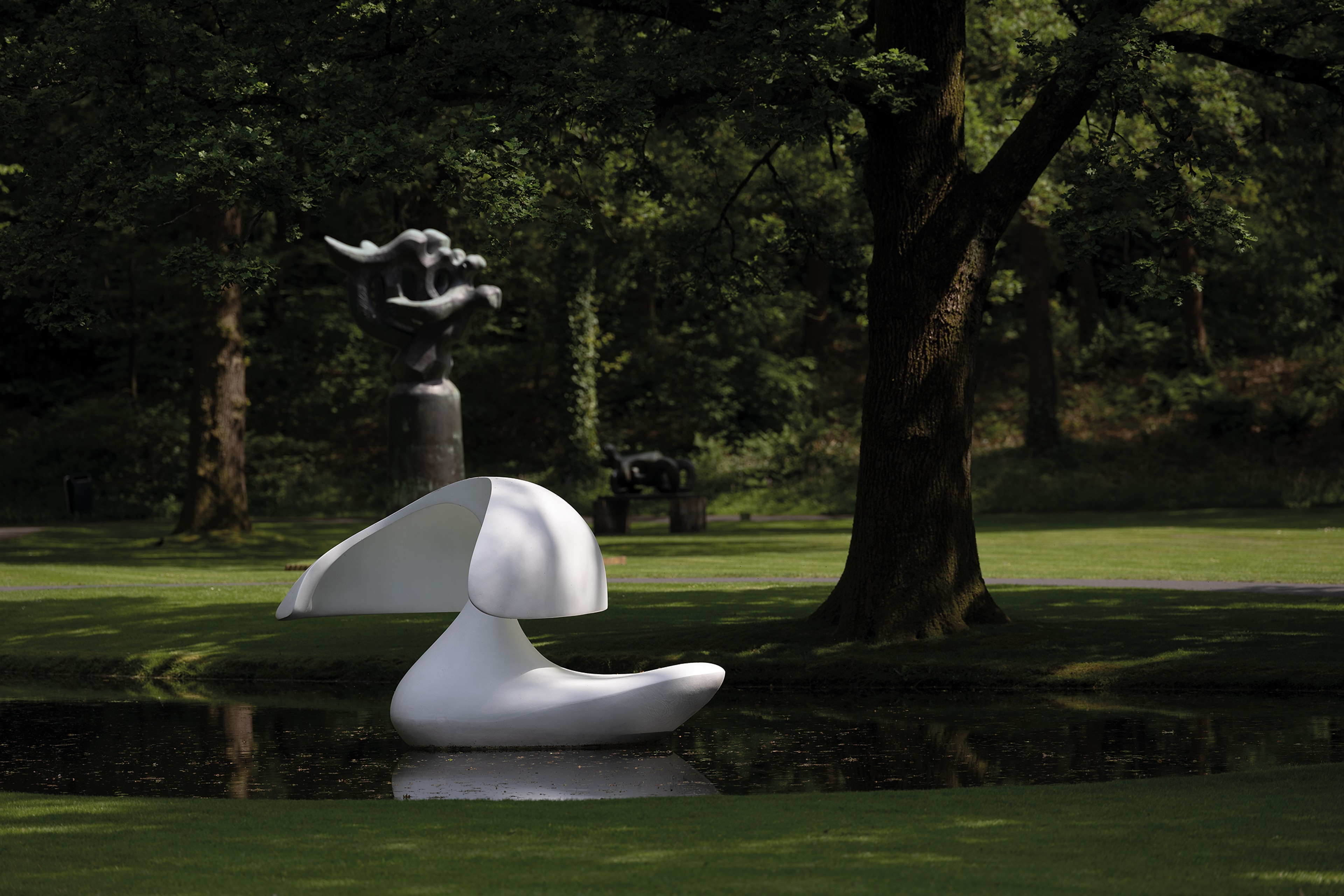 Marta Pan, _Sculpture flottante, Otterlo_, 1960-1961 Foto van een moderne, witte abstracte sculptuur geplaatst op een wateroppervlak in een parkachtige omgeving met gras en bomen. Op de achtergrond staat nog een sculptuur.