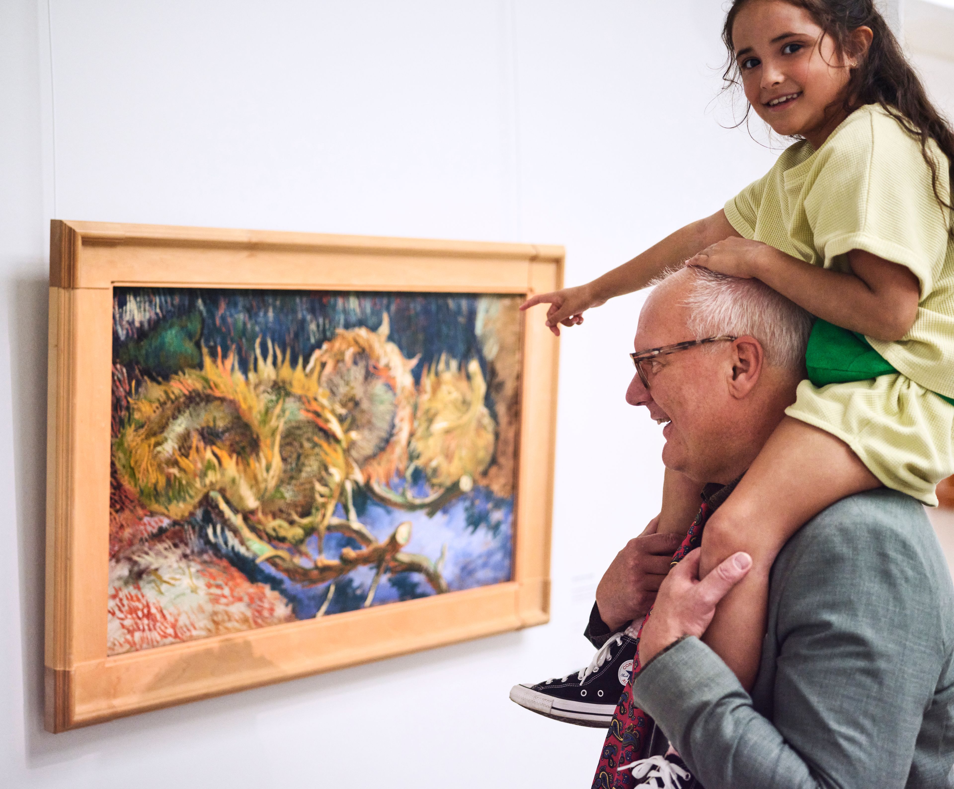 Photo of a man carrying a child on his shoulders as they look together at a colourful painting of sunflowers in a wooden frame. The scene depicts the interaction between a grandfather and his granddaughter and their appreciation of art in a museum setting.