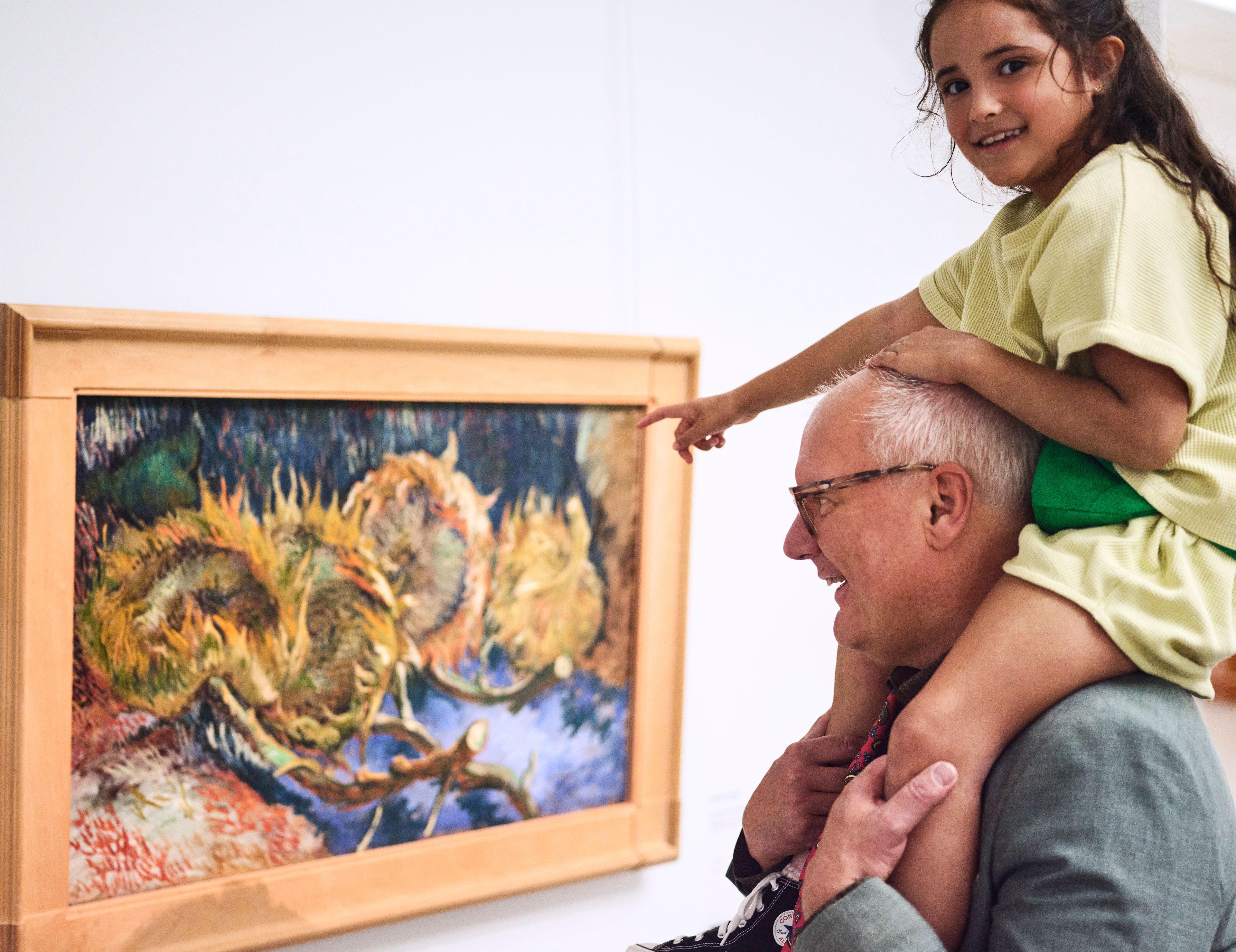 Foto van een man die een kind op zijn schouders draagt terwijl ze samen naar een kleurrijk schilderij van zonnebloemen in een houten lijst kijken. Scène toont interactie tussen opa en kleindochter en bewondering voor kunst in een museumomgeving.