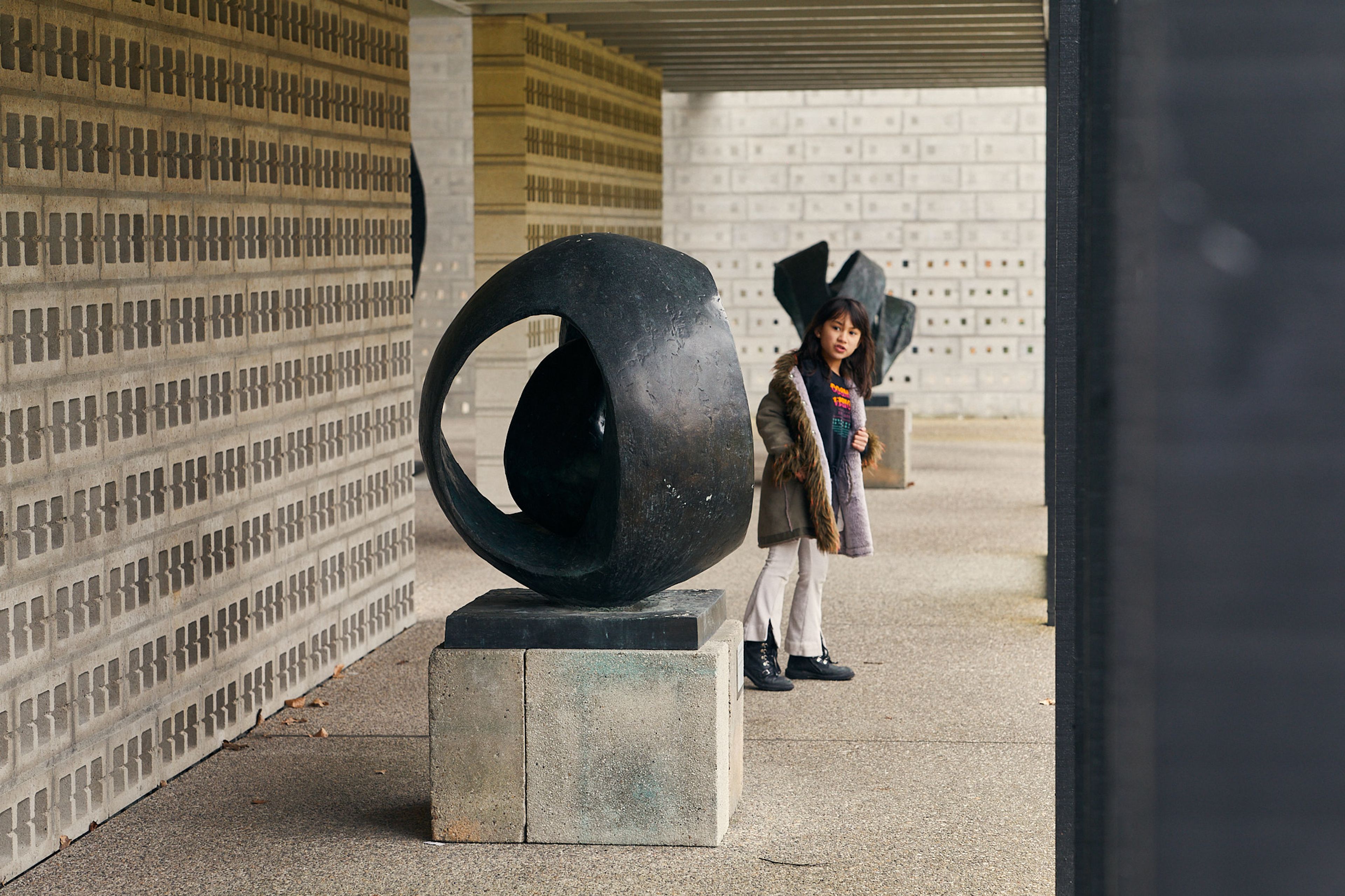 Foto van een Het Rietveldpaviljoen, met twee abstracte zwarte beelden op betonnen sokkels en een meisje dat ernaast staat. Ruimte wordt omlijst door muren met patroon van rechthoekige stenen en een betonnen vloer