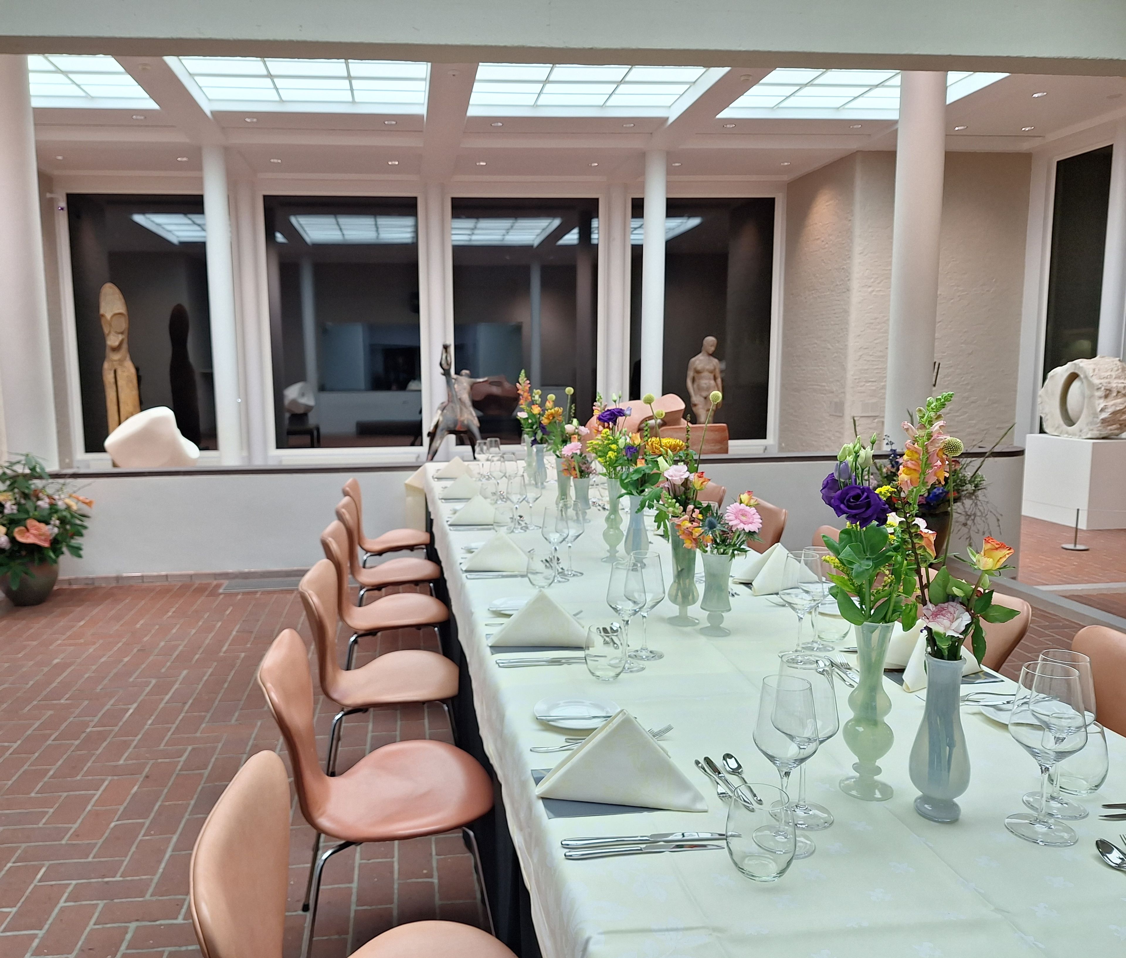 Foto van een gedekte tafel met stoelen in een beeldenzaal van het museum. Op tafel bloemen. Buiten is het donker, het is avond.