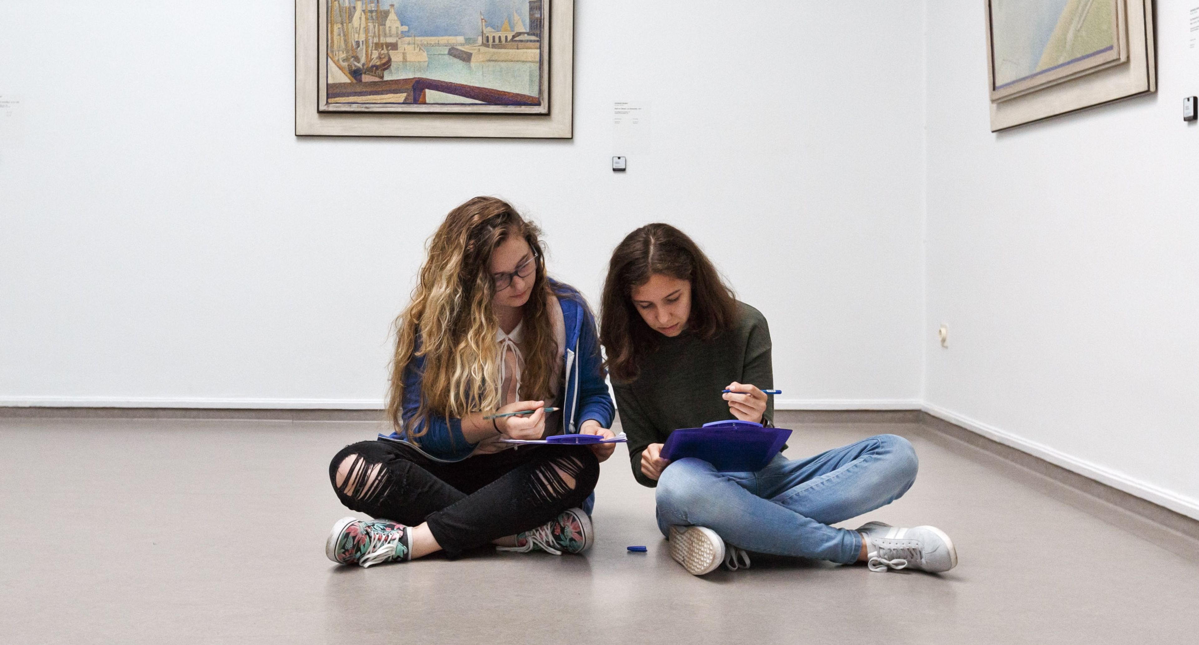 Twee zittende personen in een kunstgalerie, bezig met tekenen of schrijven op papier. Omringd door ingelijste schilderijen aan witte muren, toont scène een rustige, creatieve omgeving.