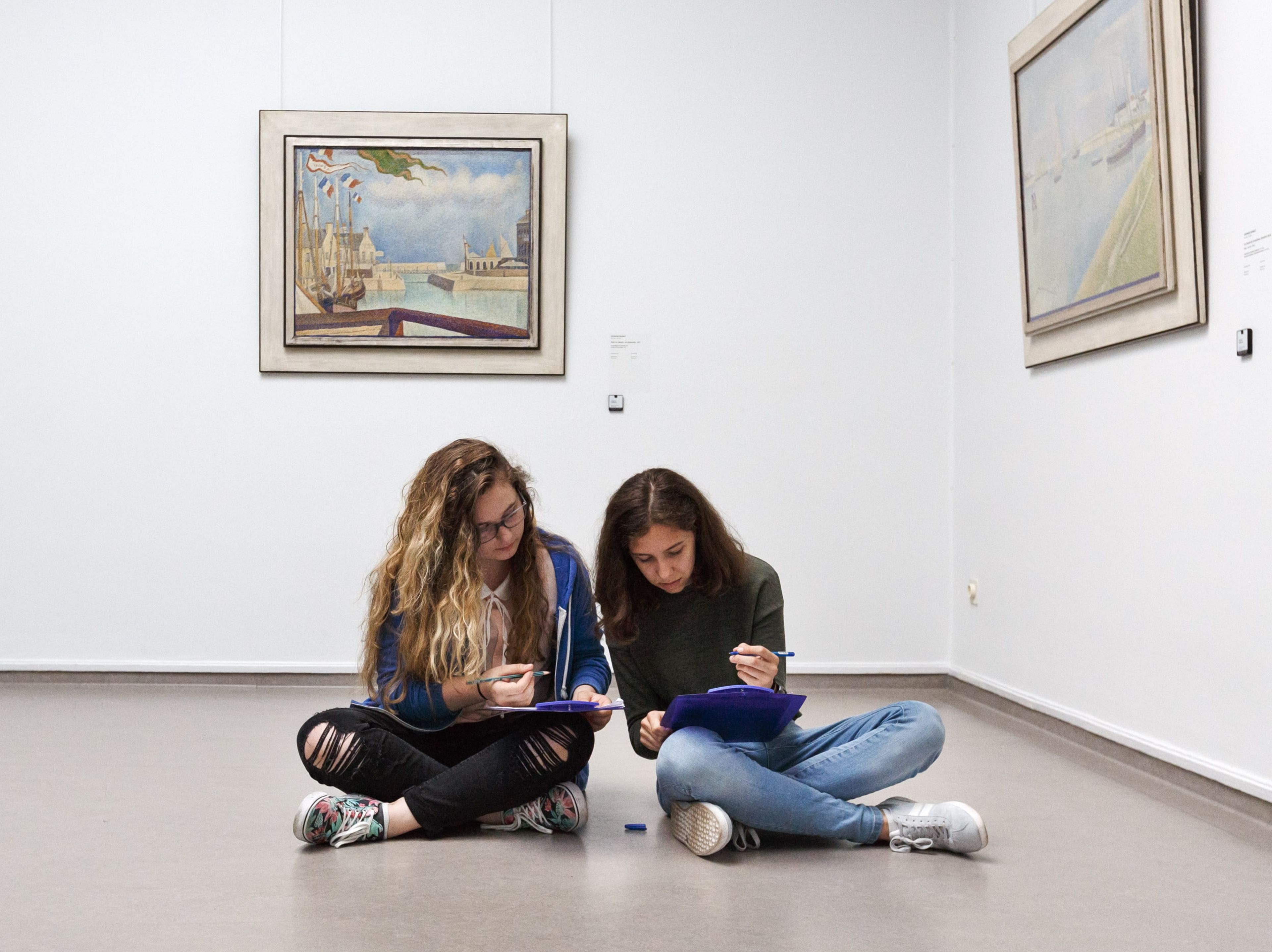Twee zittende personen in een kunstgalerie, bezig met tekenen of schrijven op papier. Omringd door ingelijste schilderijen aan witte muren, toont scène een rustige, creatieve omgeving.